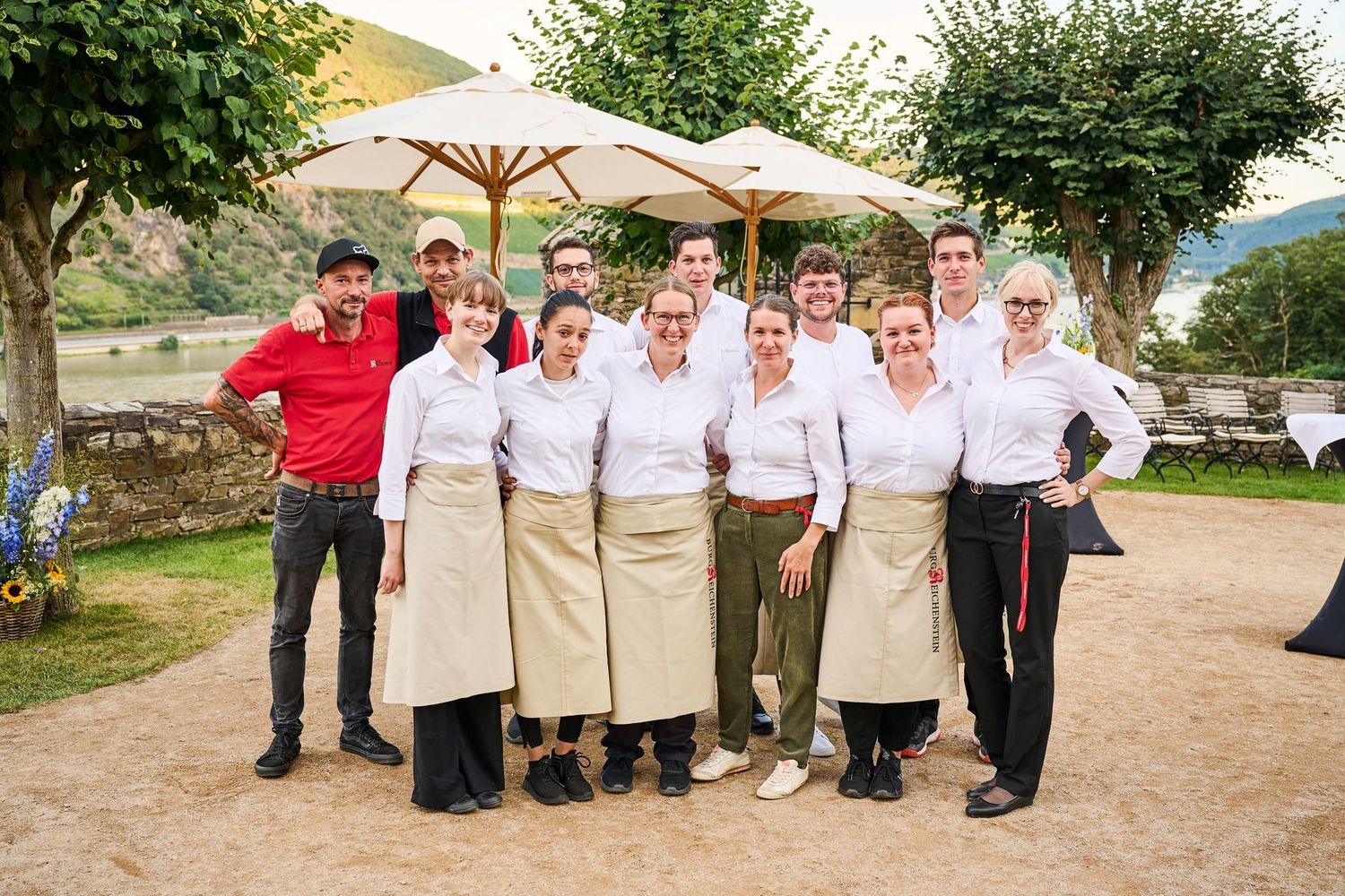 Eine Gruppe Restaurantmitarbeiter posiert im Freien in weißen Hemden und beigen Schürzen und lächelt vor Sonnenschirmen und Bäumen.
