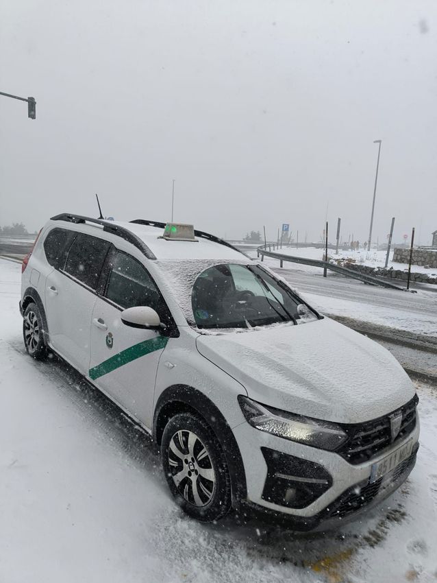 Taxi blanco cubierto de nieve, estacionado en una calle nevada. Franja verde en el lateral. Cielo nublado.