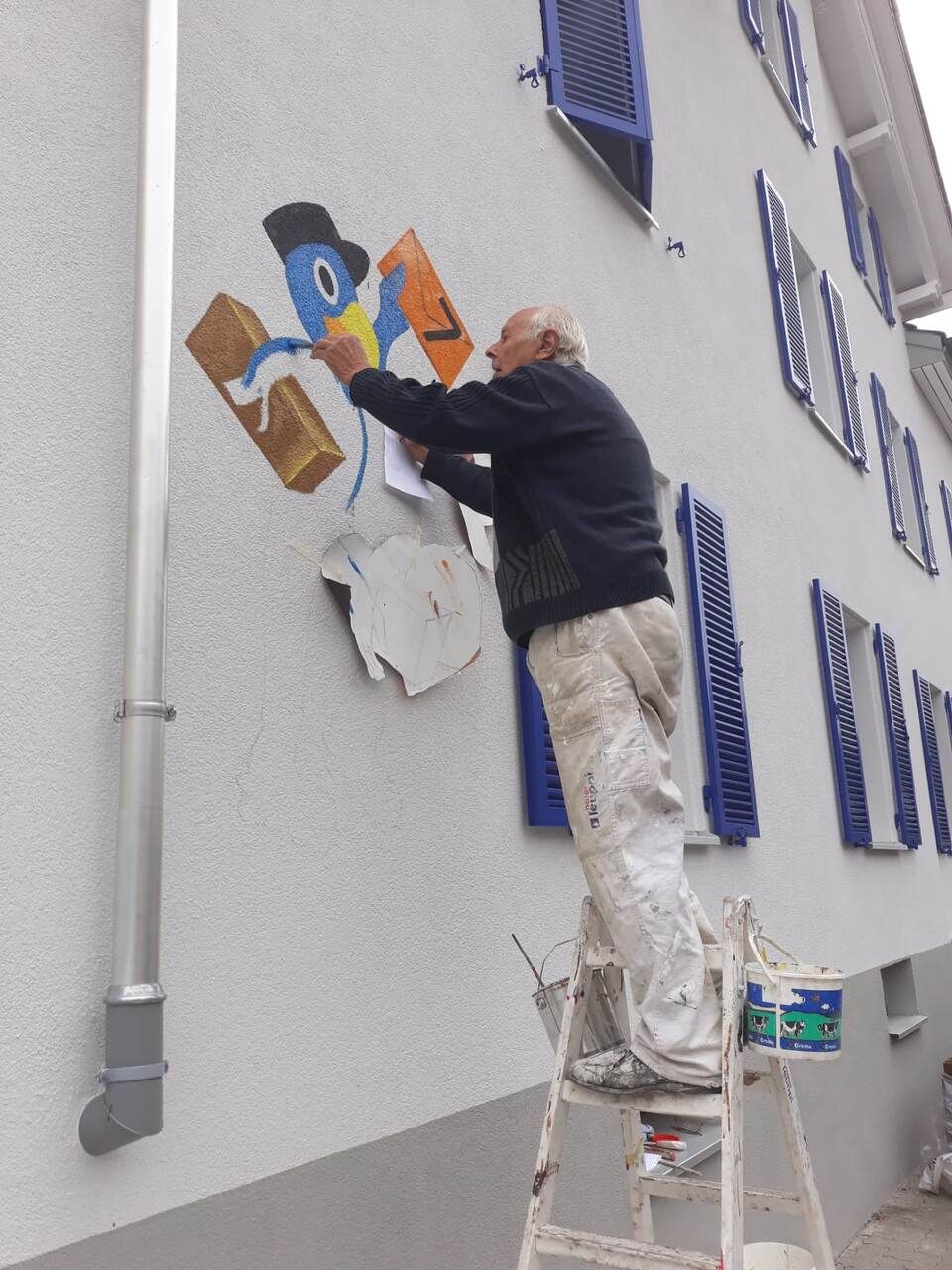 Ein Mann auf einer Leiter malt ein Bild an die Seite eines Gebäudes. Foto von der Maler Leuppi GmbH