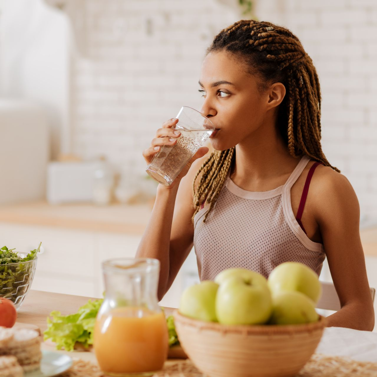 Une femme boit de l'eau à une table garnie de fruits, de légumes et de jus.