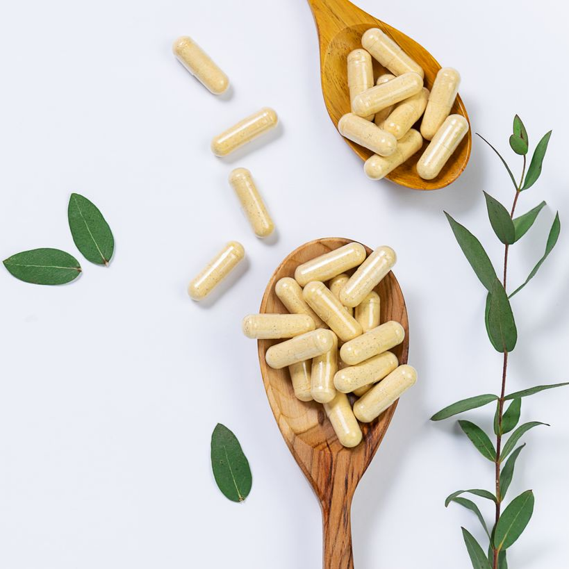 Capsules beiges dans des cuillères en bois avec des feuilles vertes sur un fond blanc.