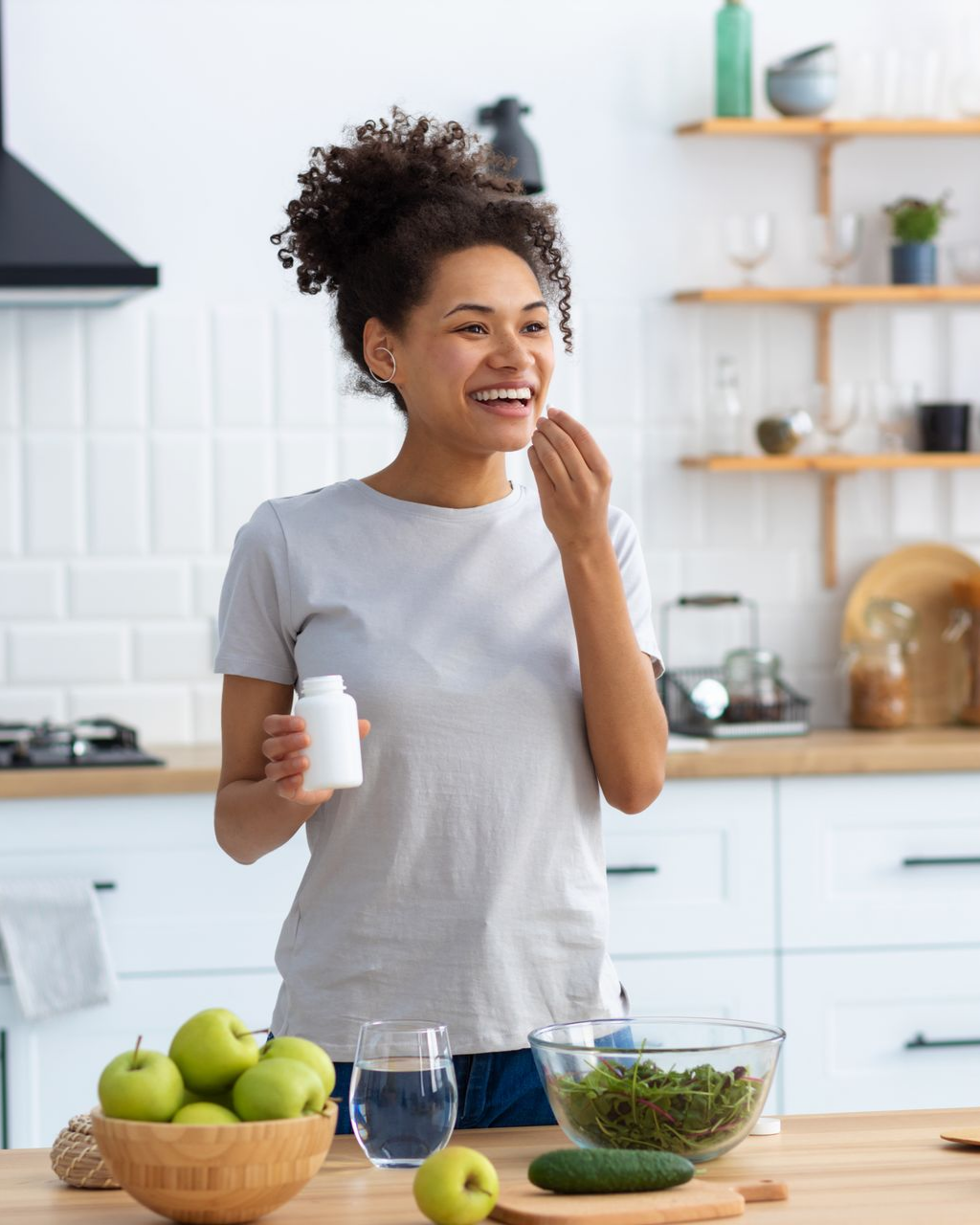 Une femme souriante prend un comprimé dans un flacon blanc, dans sa cuisine.