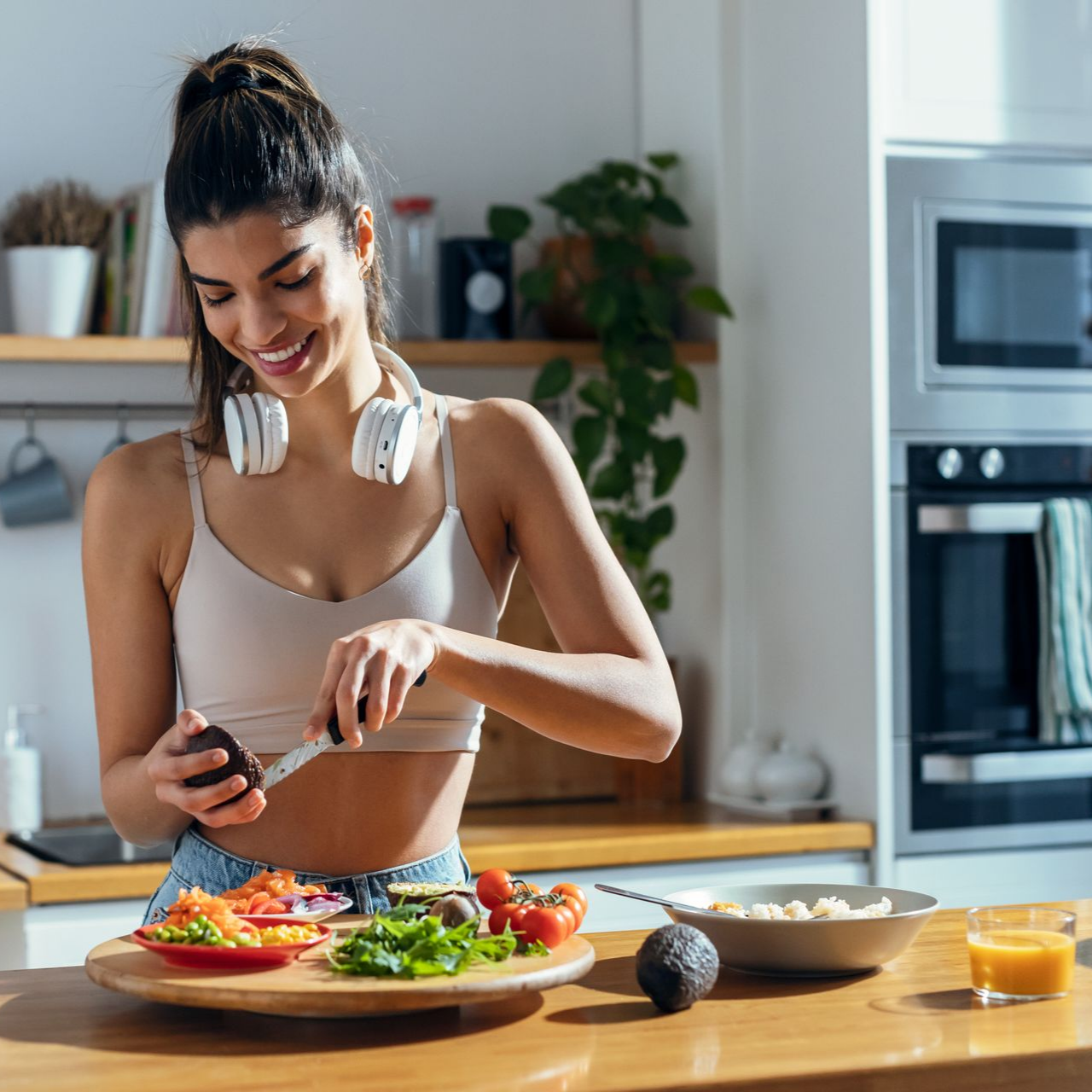 Une femme souriante, écouteurs autour du cou, coupe un avocat dans une cuisine.