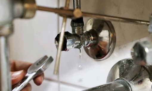 Nahaufnahme eines tropfenden Badezimmerwasserhahns, aus dem Wasser in ein Waschbecken tropft; daneben hält eine Hand einen Schraubenschlüssel.