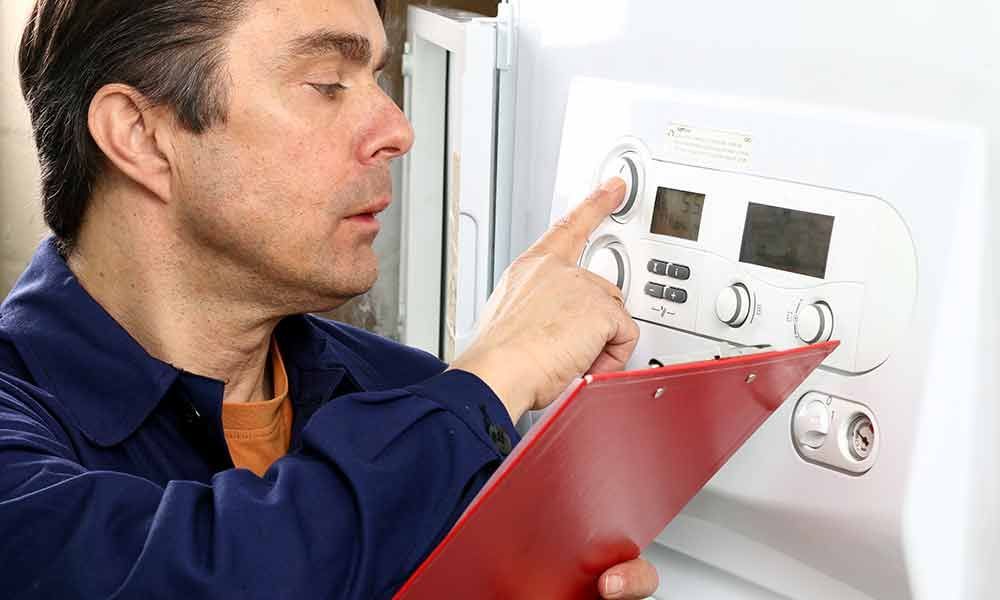 Ein Techniker im blauen Hemd überprüft einen Wandthermostat und dessen Bedienelemente, während er ein rotes Klemmbrett hält.