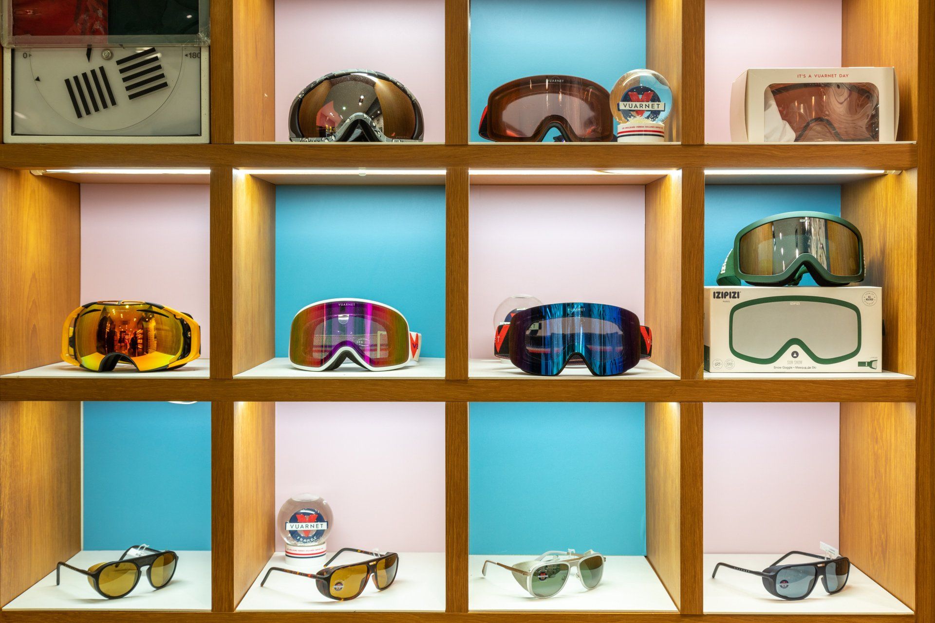 Présentation de lunettes de ski et de lunettes de soleil sur une étagère en bois à fond bleu et rose.