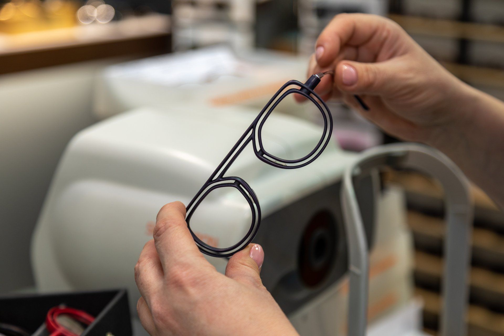 Des mains tenant des montures de lunettes noires dans un magasin d'optique, une machine en arrière-plan.