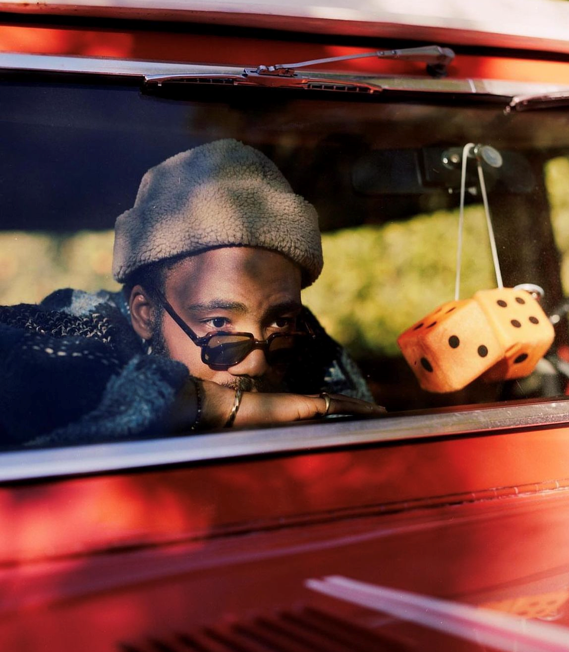 Une personne appuyée contre la vitre d'une voiture rouge, portant des lunettes de soleil et une casquette. Des dés en peluche sont accrochés à proximité.