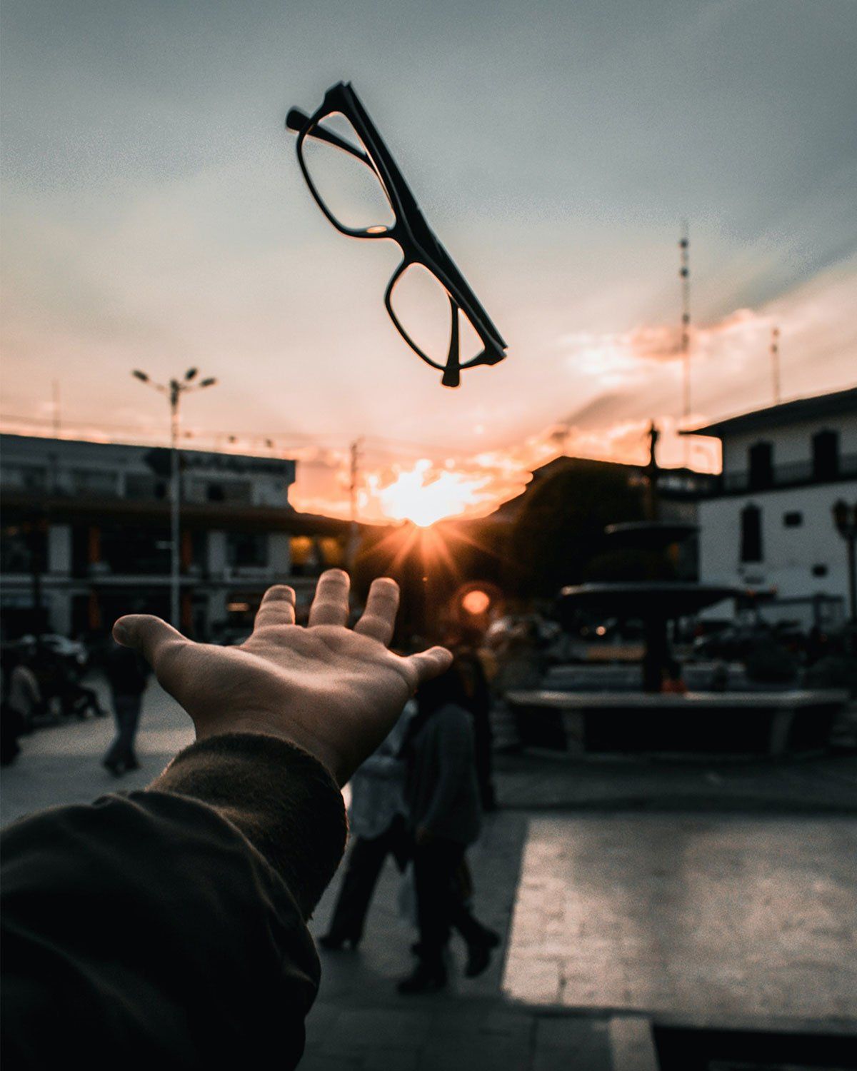 Une main jette des lunettes en l'air sur fond de coucher de soleil au-dessus d'une place de ville.