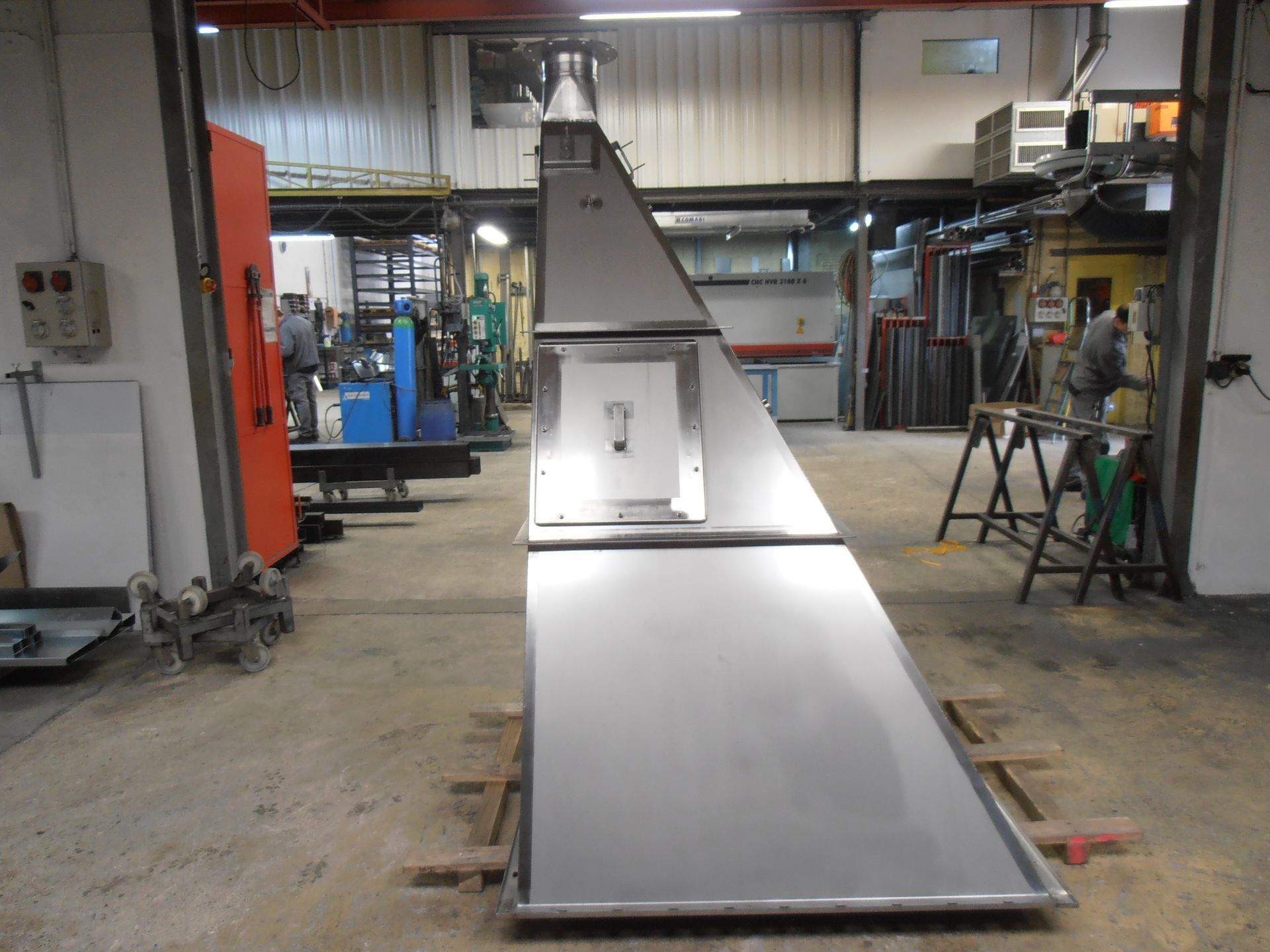 Grande structure métallique argentée et angulaire dans un atelier ; une base rectangulaire se rétrécit vers le haut pour former une cheminée.