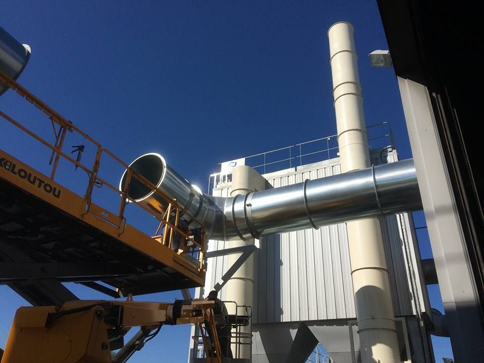 Un ouvrier sur un ascenseur installe un conduit métallique fixé à une haute structure industrielle blanche contre un ciel bleu.