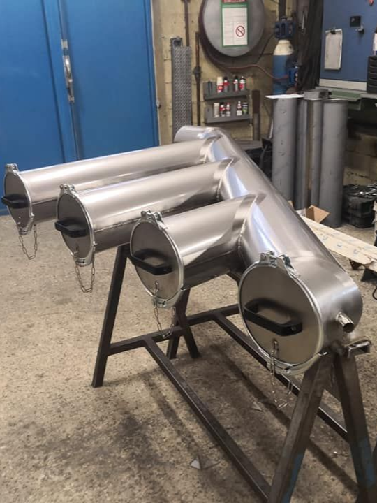 Collecteur d'échappement en métal argenté à quatre chambres latérales, reposant sur un support métallique dans un atelier.