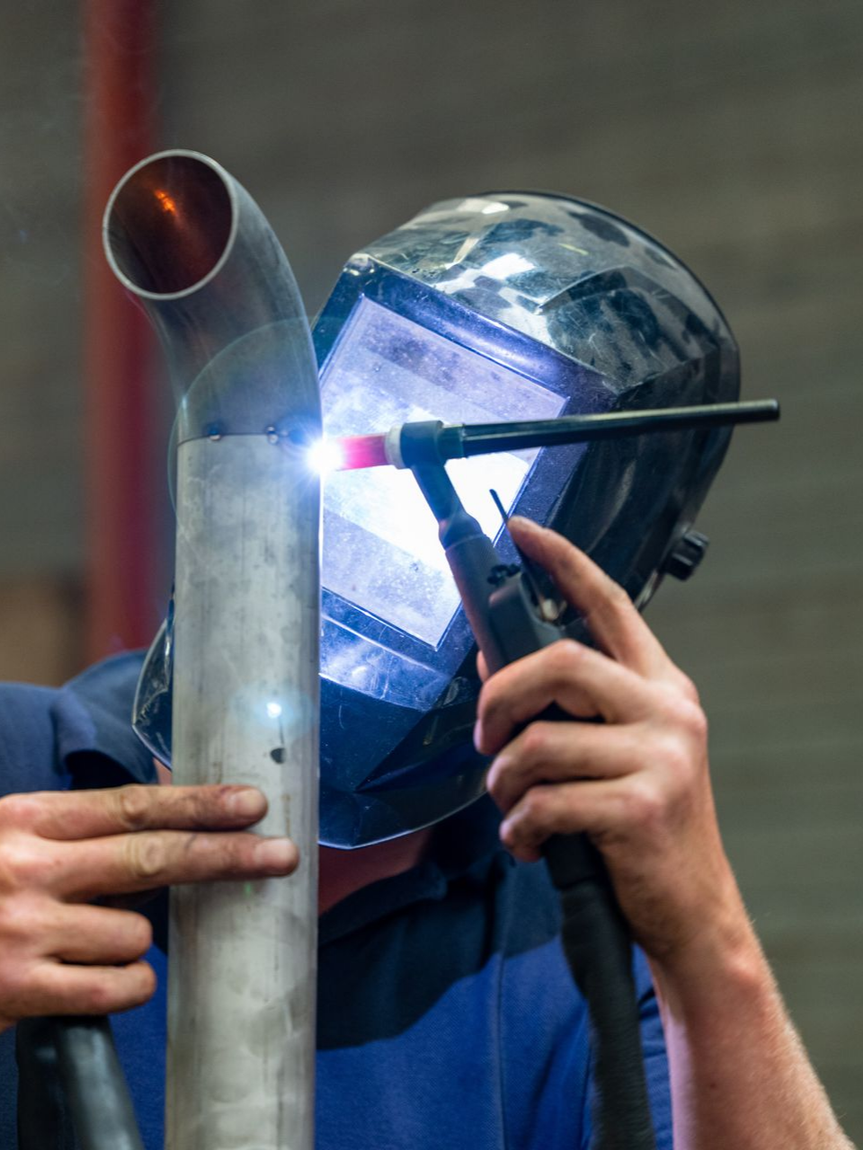 Soudeur en chemise bleue utilisant un chalumeau sur un tuyau métallique dans un atelier.