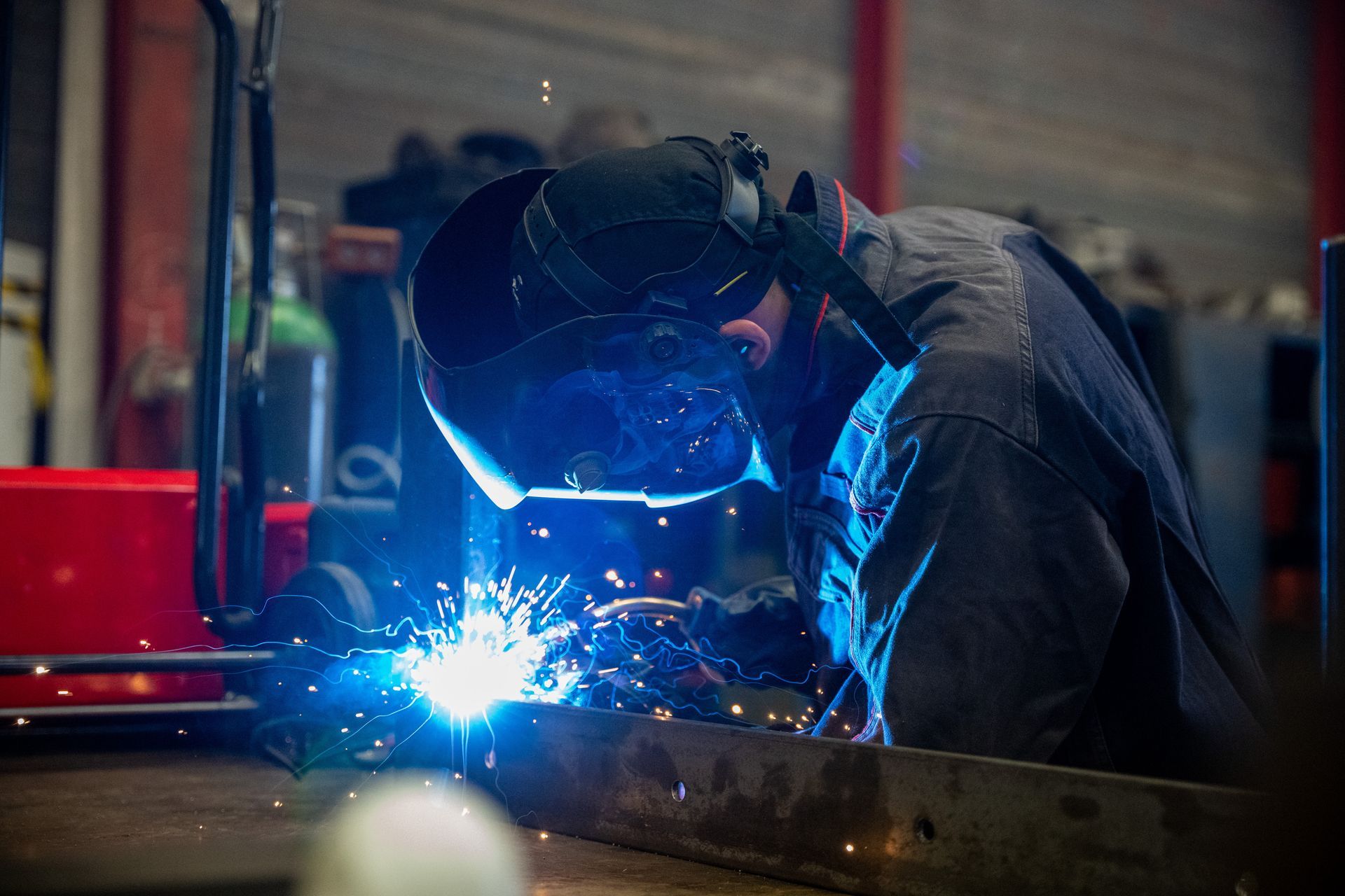 Soudeur en salopette sombre avec un casque, des étincelles jaillissent, travaillant sur du métal dans un atelier.