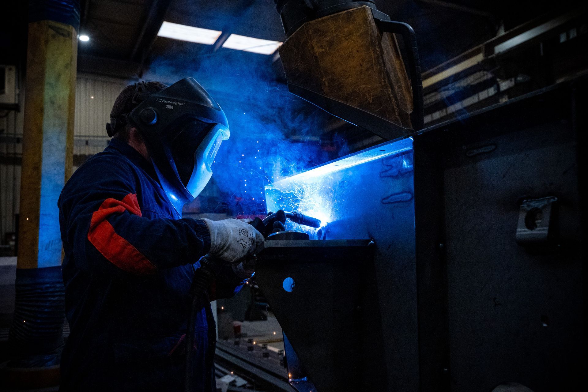 Soudeur en tenue de protection bleu foncé, soudant avec des étincelles et une lumière bleu vif dans un atelier de métallurgie.
