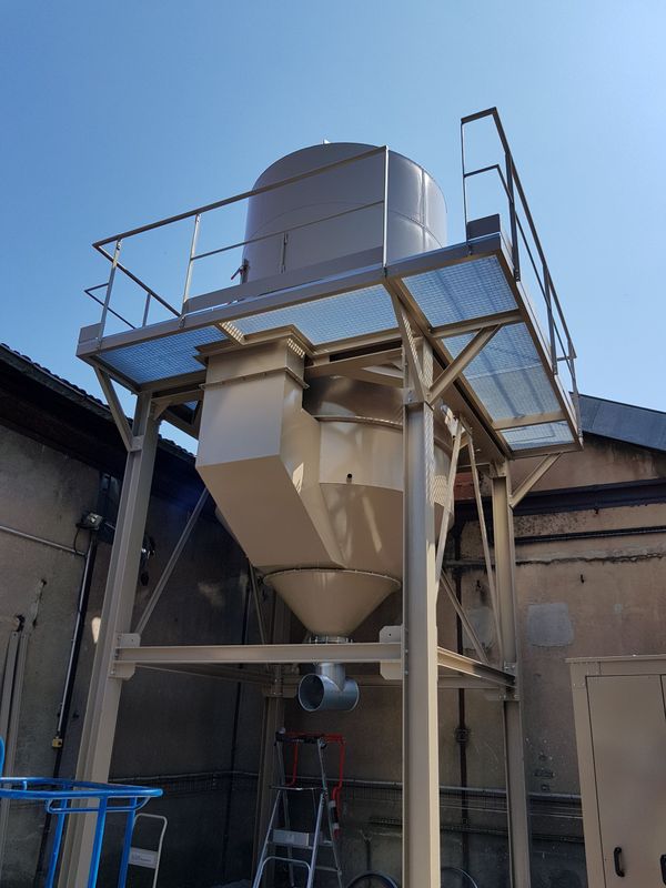 Silo industriel en métal sur une plate-forme avec garde-corps, à l'extérieur contre un bâtiment et un ciel bleu.