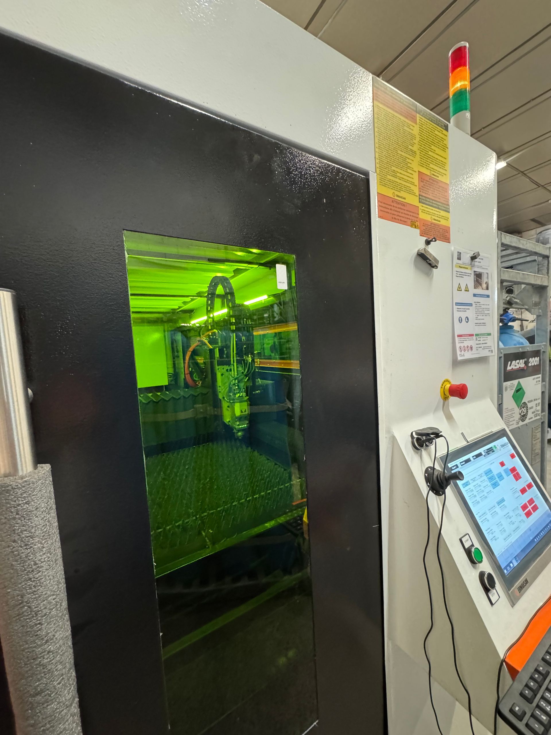 Machine de découpe laser avec intérieur éclairé en vert visible à travers une fenêtre rectangulaire. Panneau de commande et éclairage de sécurité sur le côté.
