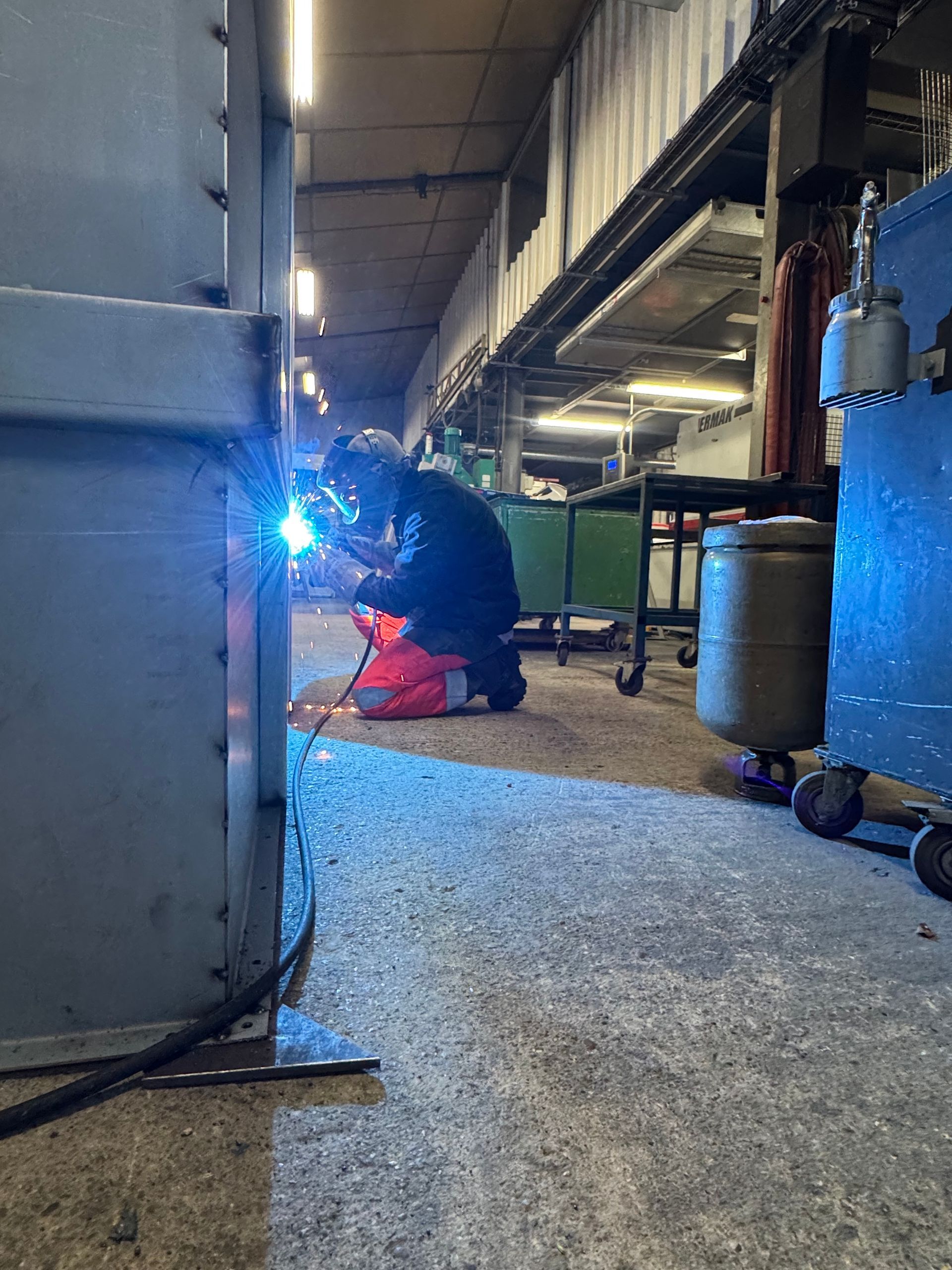 Soudeur en tenue de protection, agenouillé, en train de souder du métal dans un atelier, arc électrique brillant, lumière bleue.