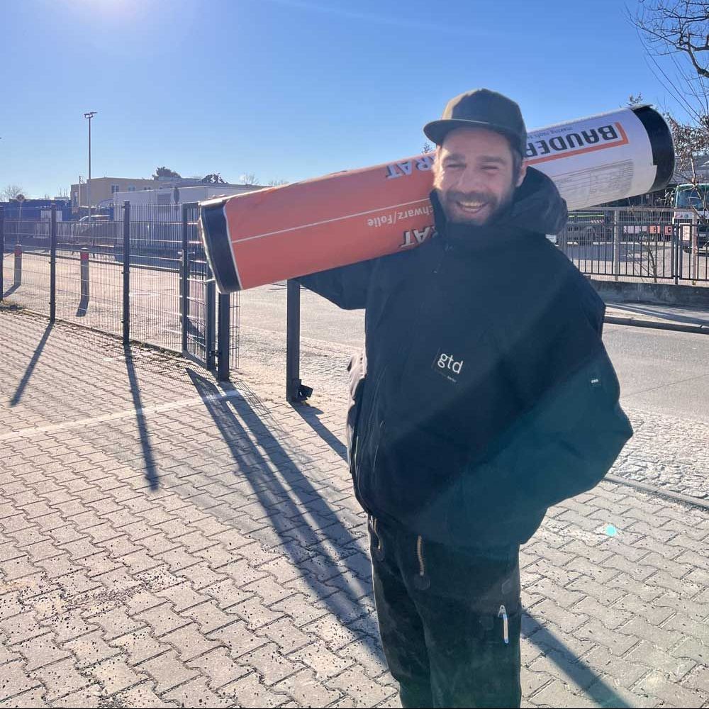 Ein Bauarbeiter in Overall und Schutzhelm lächelt in die Kamera.