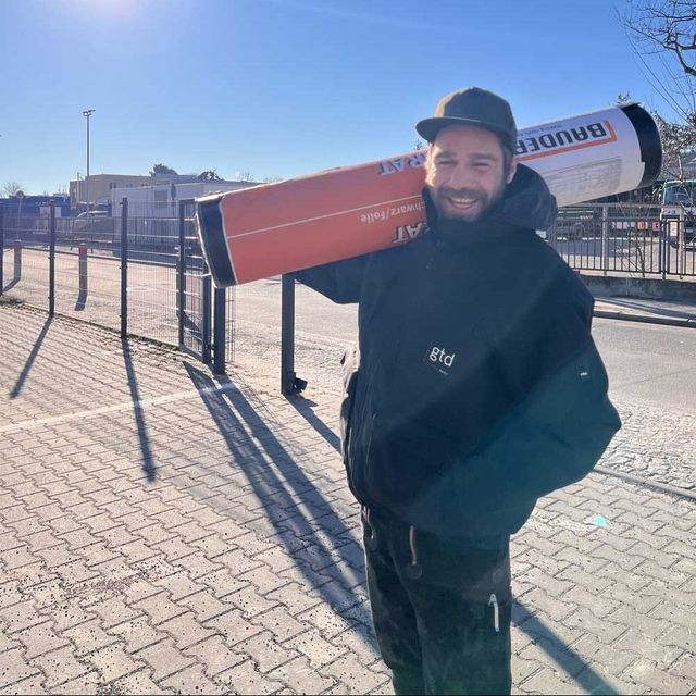 Ein Bauarbeiter in Overall und Schutzhelm lächelt in die Kamera.