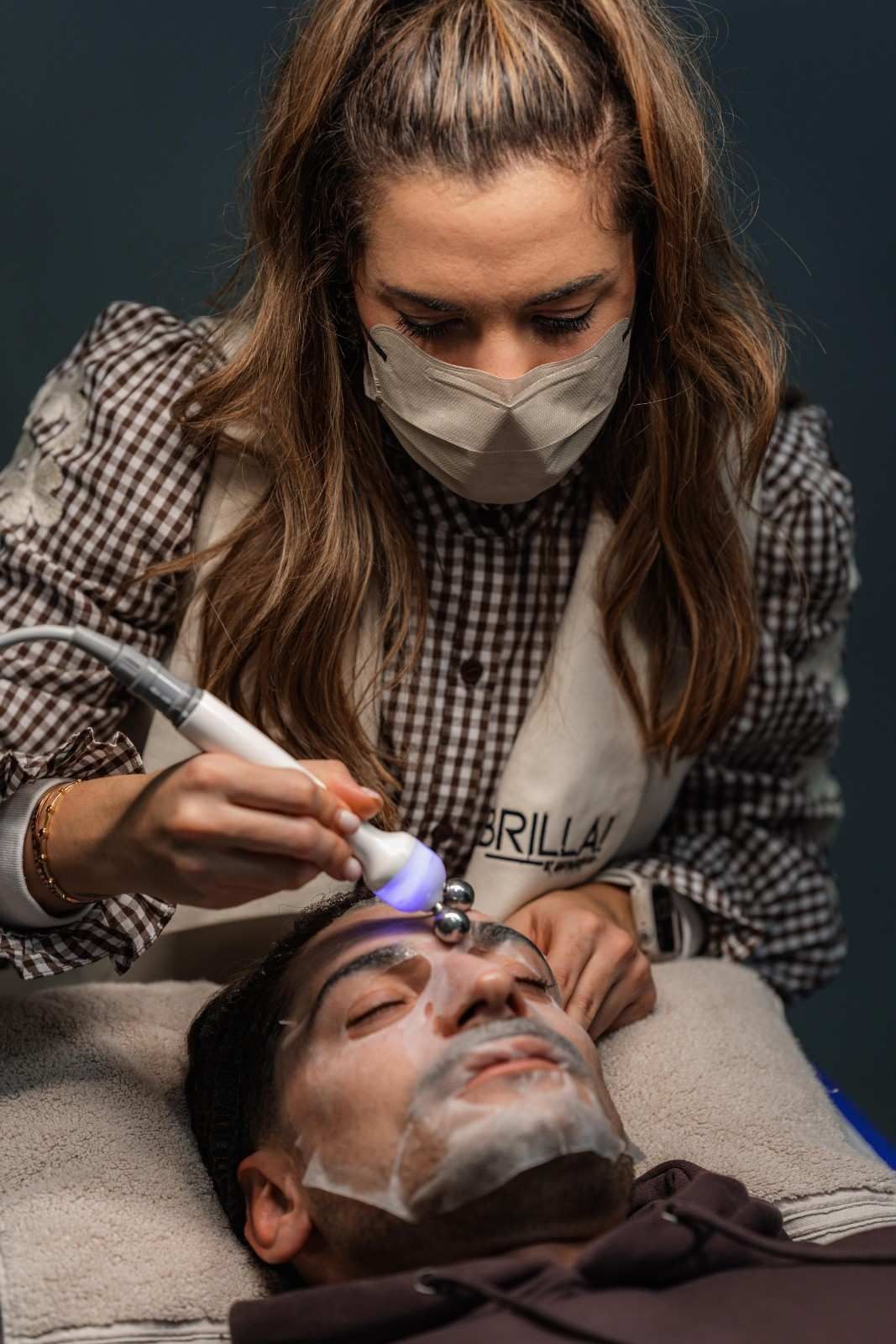 Una persona recibe un tratamiento facial de un esteticista enmascarado, utilizando un dispositivo portátil en un salón de belleza.