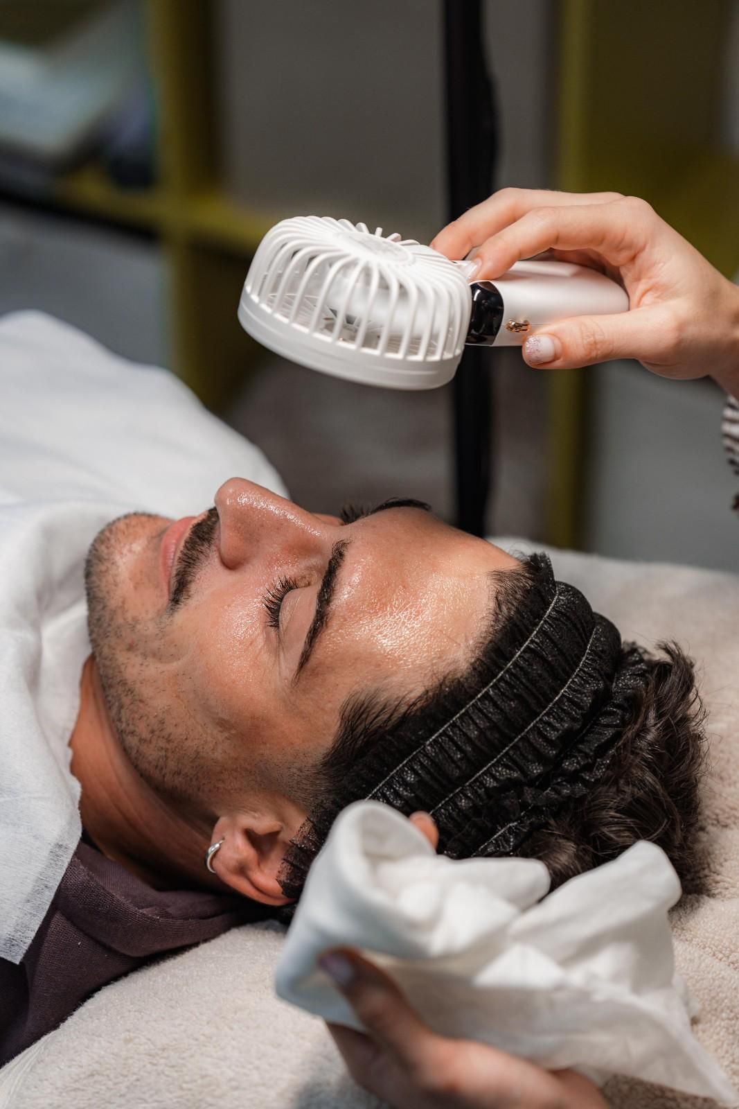 Persona recibiendo tratamiento facial; ventilador sobre la cara y toalla en la mano.