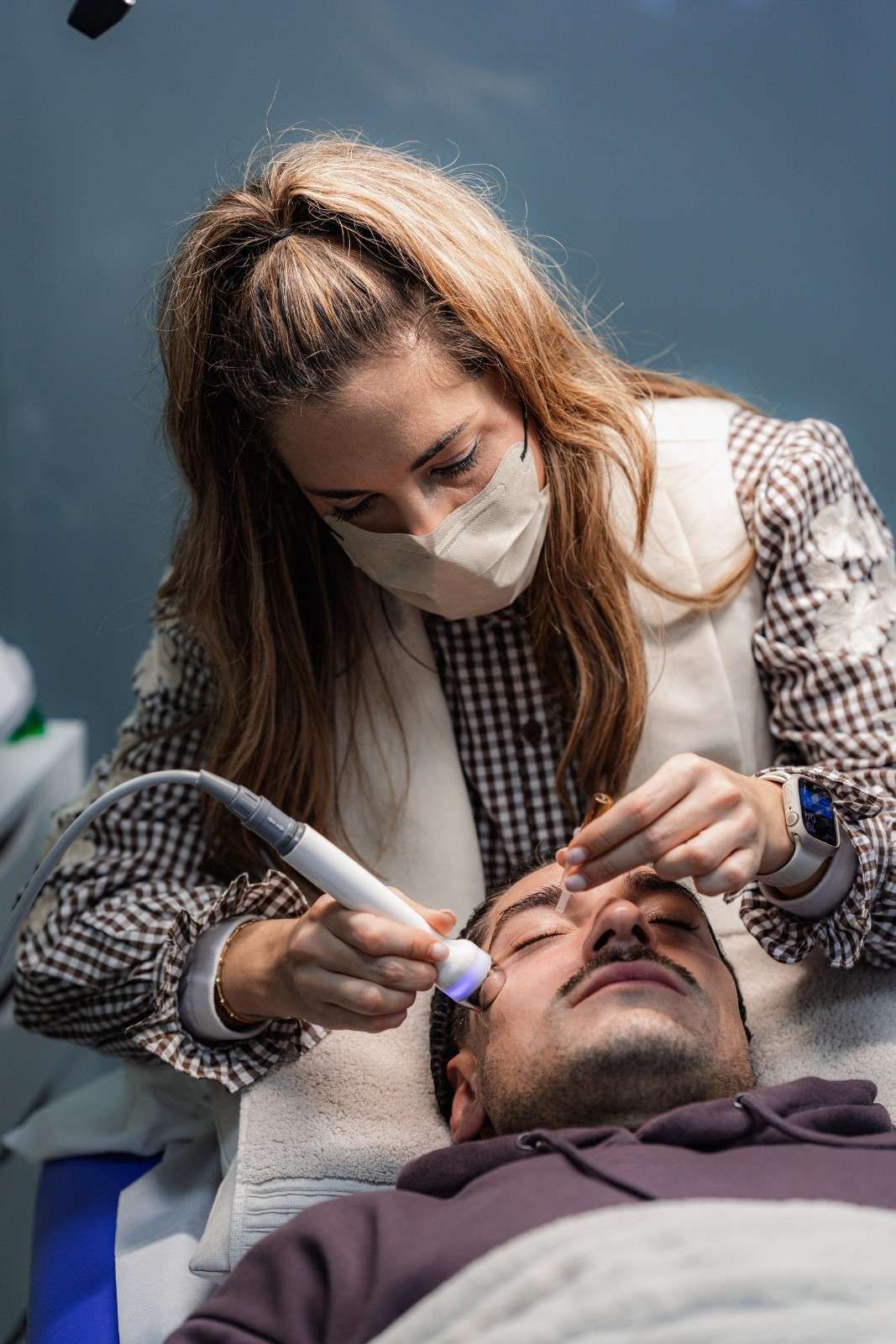 Mujer con máscara realiza un tratamiento facial a una persona acostada.