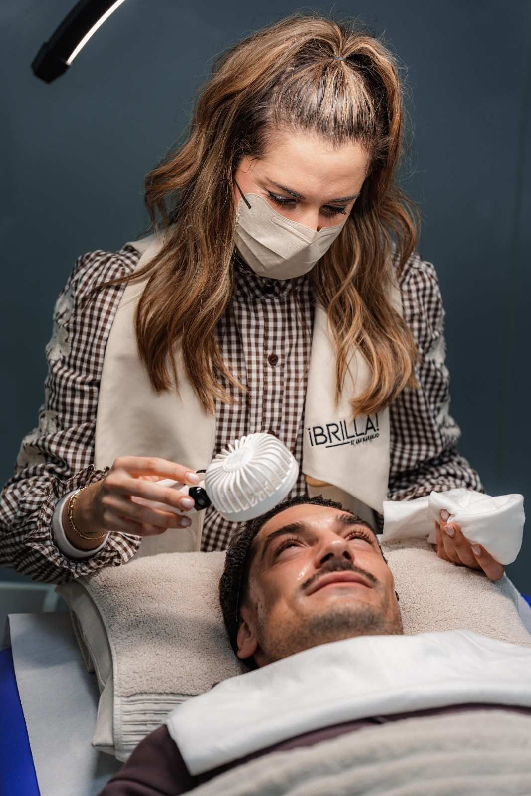 Una mujer con mascarilla trata la cabeza de un paciente. Sostiene un dispositivo cerca de su rostro en un entorno médico.