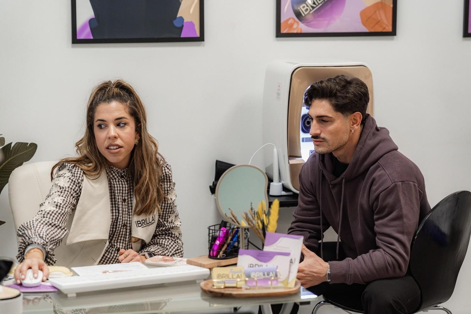 Mujer sentada en un escritorio, gesticulando, hablando con un hombre sentado frente a ella. Ambiente de oficina, colores pastel.