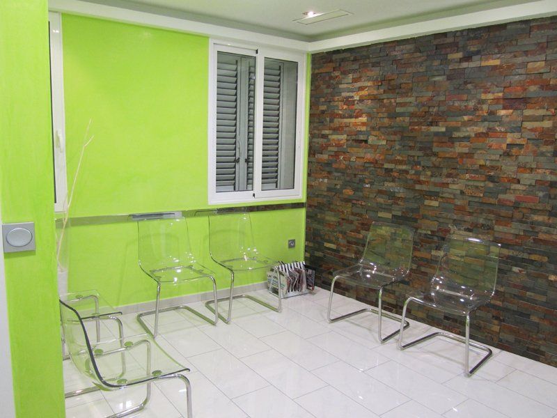 Habitación vacía con paredes de color verde lima y piedra, suelo de baldosas blancas y sillas transparentes cerca de una ventana.