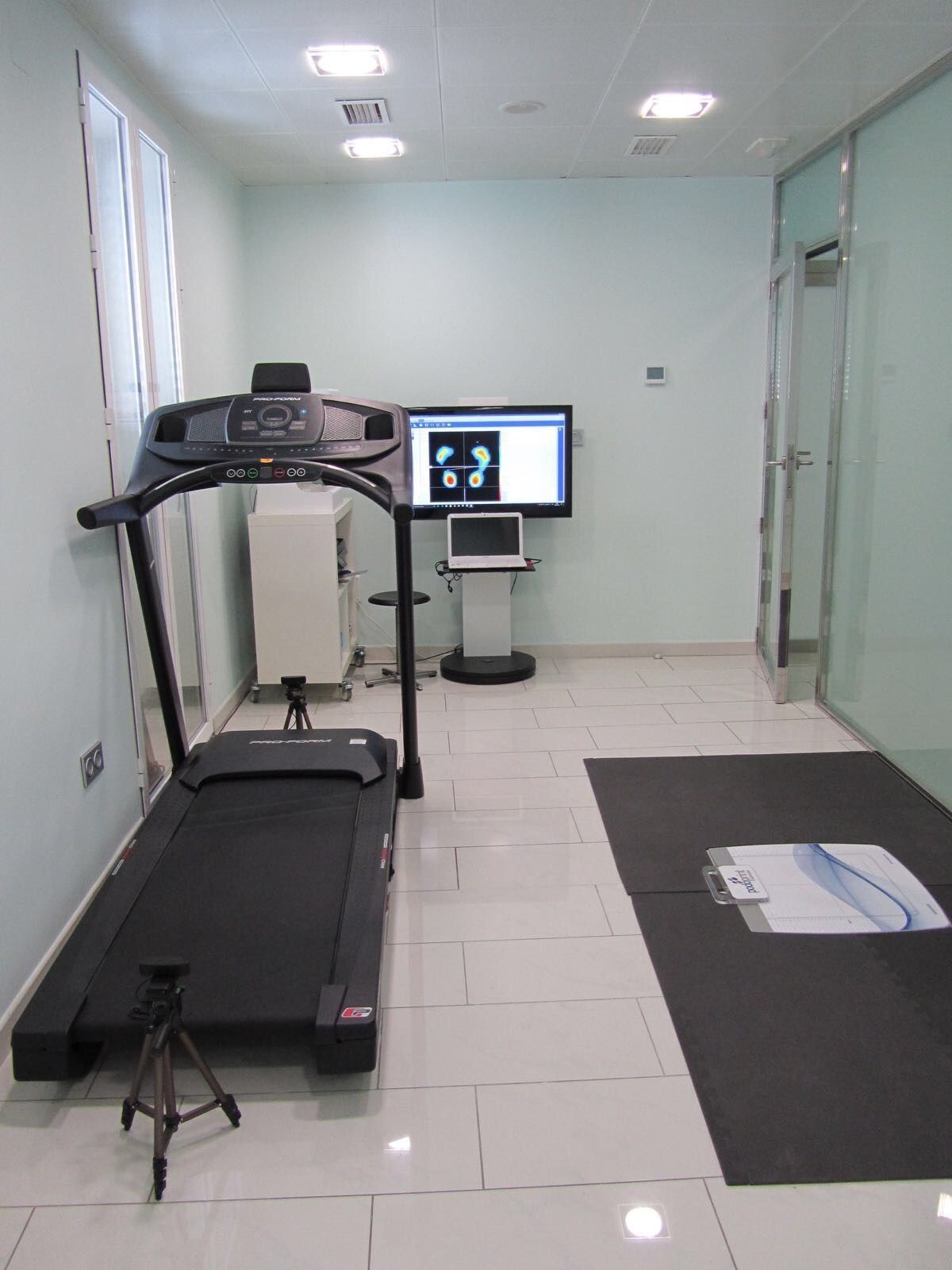Habitación de clínica vacía con una cinta de correr y un monitor sobre un soporte cerca de la pared del fondo.