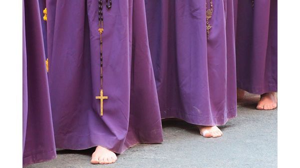 Pies de personas con túnicas moradas de pie sobre un suelo de color claro, con un collar de cruz dorada colgando.