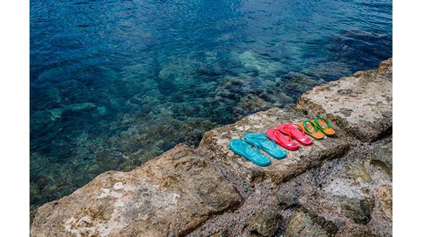 Chanclas de colores alineadas en una orilla rocosa junto a aguas cristalinas de color azul.