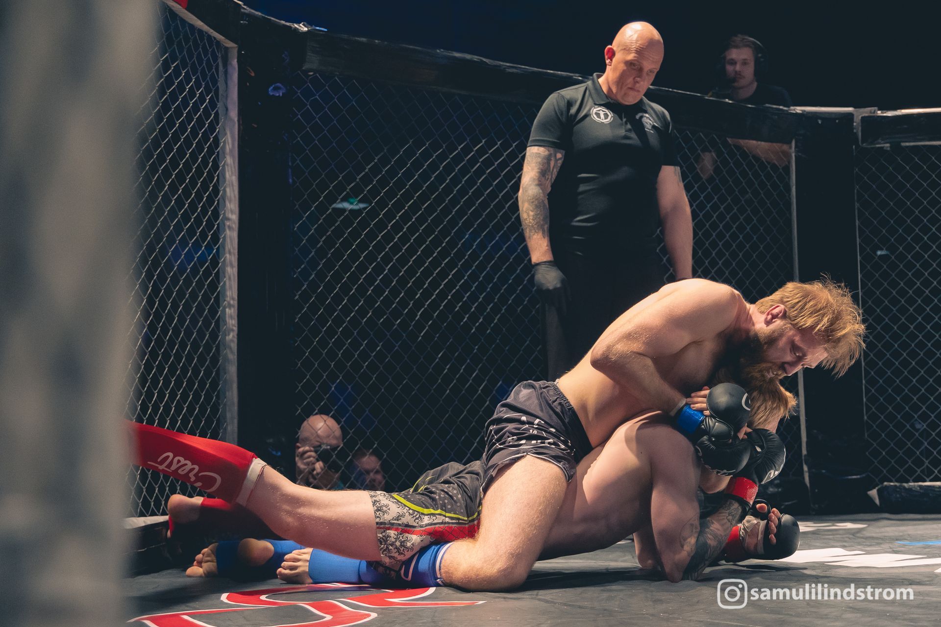MMA-ottelu häkissä, yksi ottelija painaa toista matolle tuomarin katsellessa lähellä