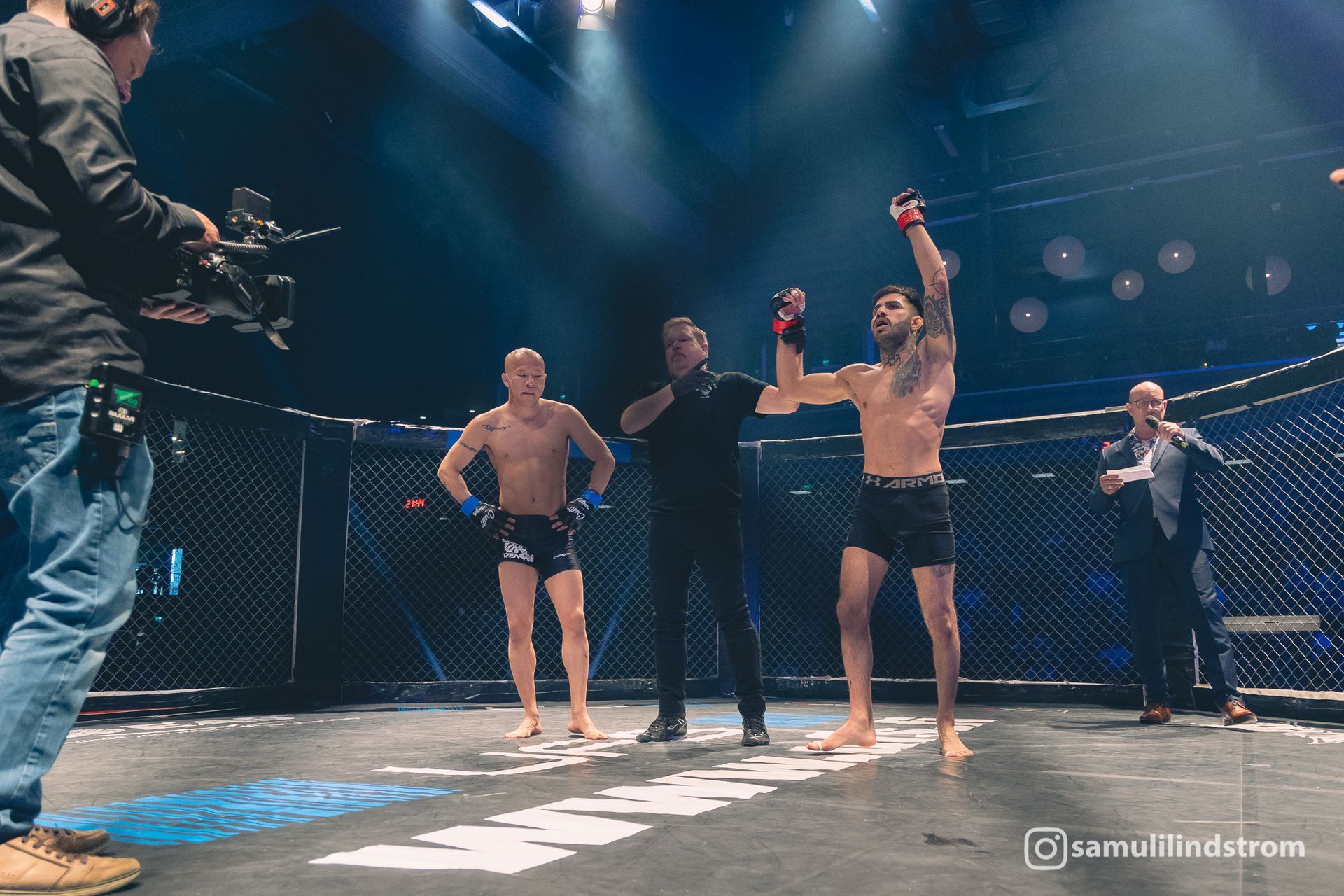 MMA-ottelijat ja tuomari kahdeksankulmiossa, toinen ottelija nostaa kätensä voiton merkiksi areenan valojen alla