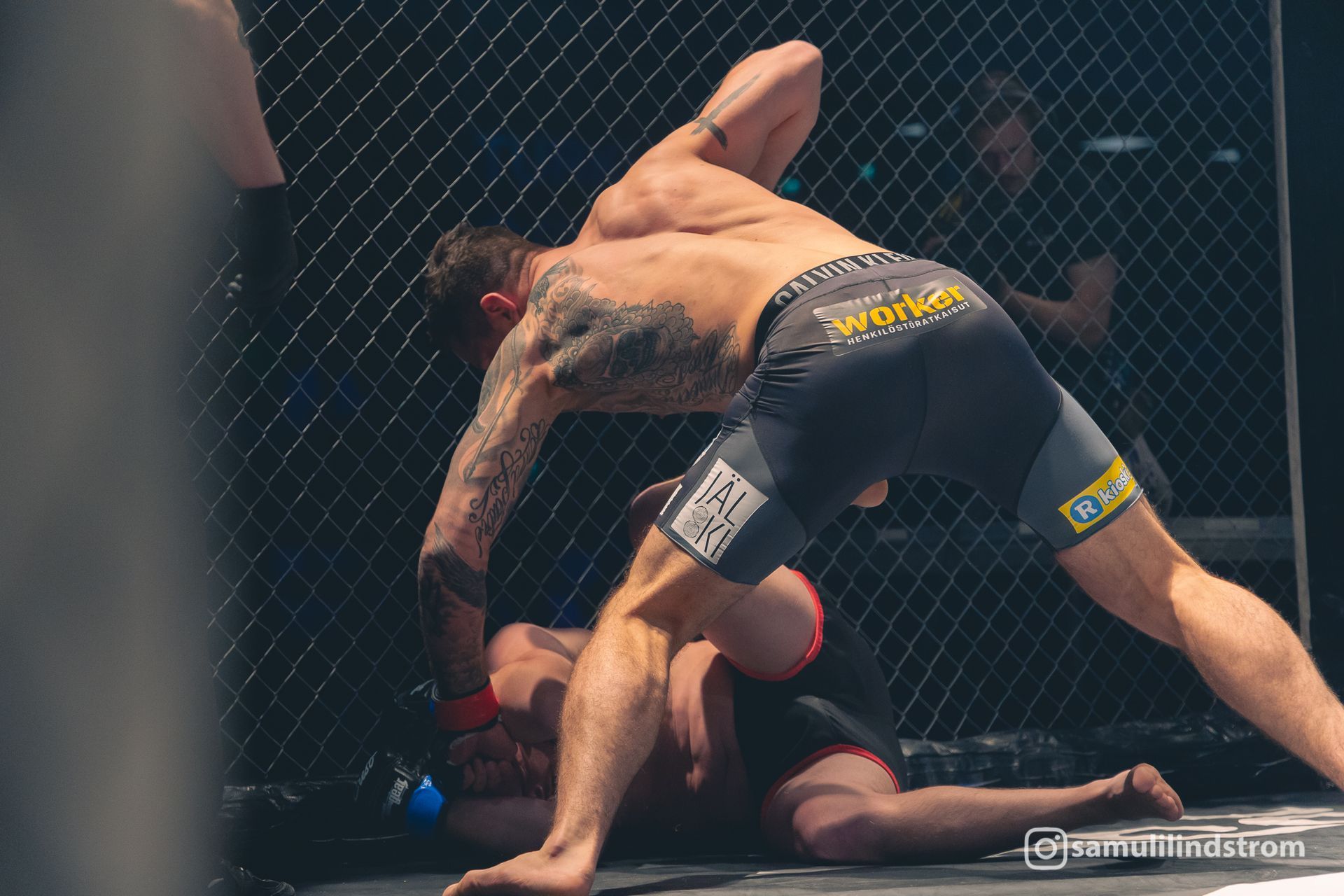 MMA-ottelija mustissa shortseissa hakkaa vastustajaa häkissä ottelun aikana