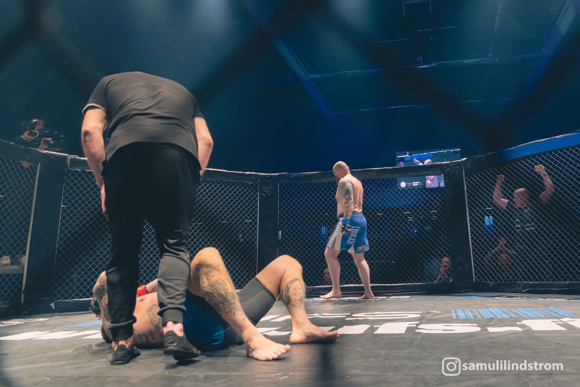 MMA-ottelu häkissä: yksi ottelija on matolla, toinen seisoo lähellä ja tuomari nojaa heidän ylleen.