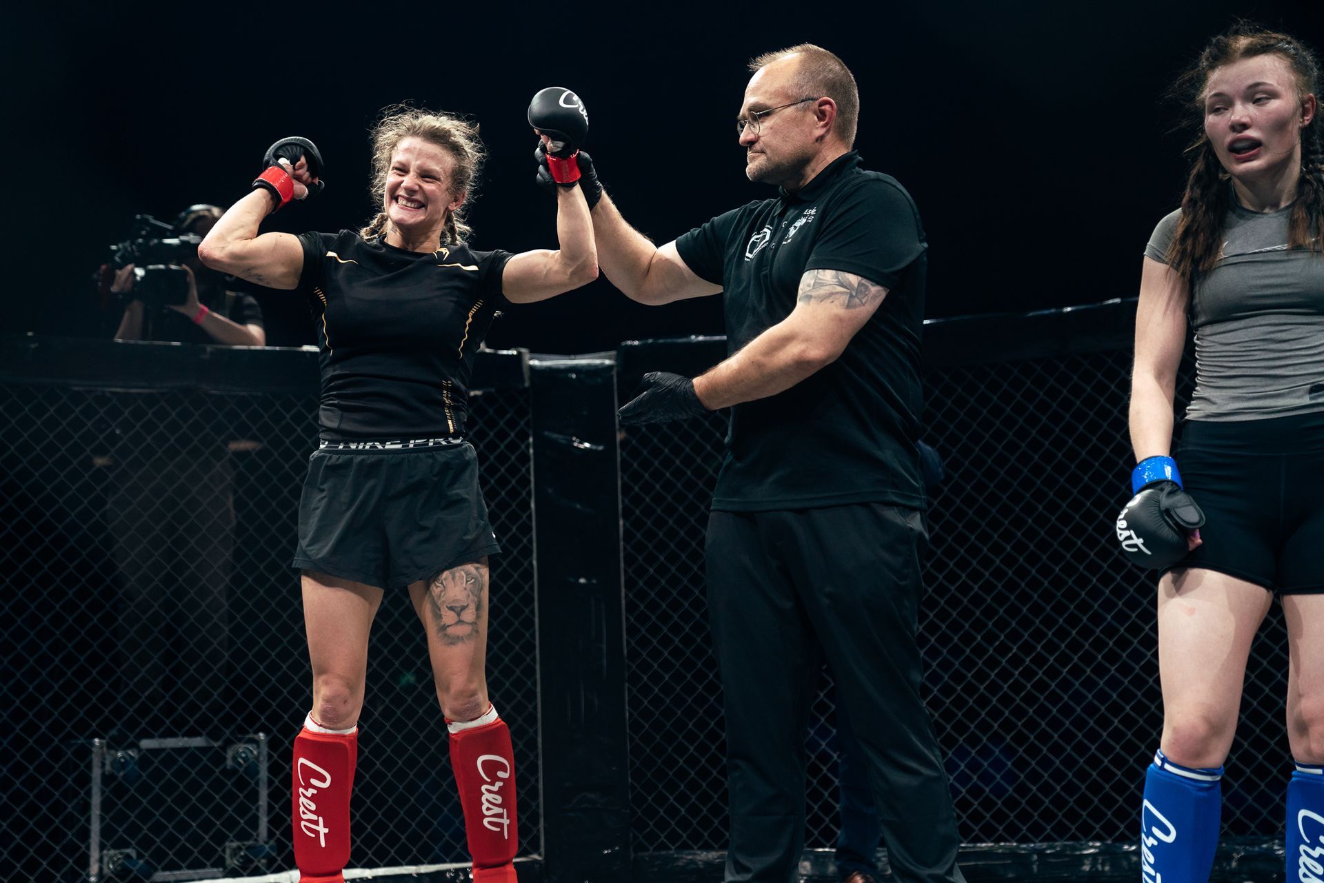 MMA-tuomari nostaa ottelijan käden häkissä, kun toinen ottelija seisoo lähellä ottelun jälkeen.