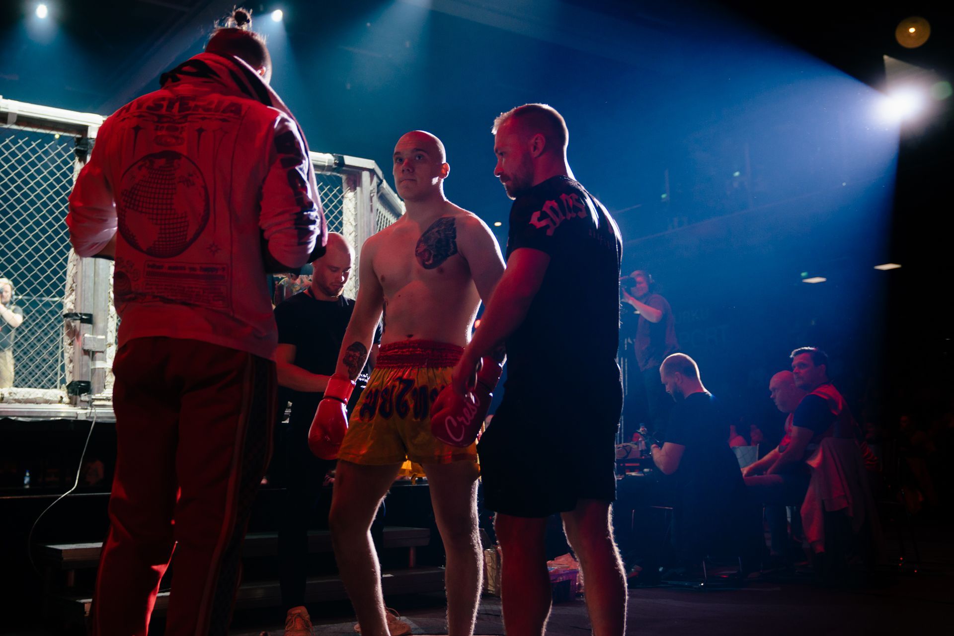 MMA-ottelijat ja tuomari kehässä kirkkaiden areenavaloissa, taustalla katsojia