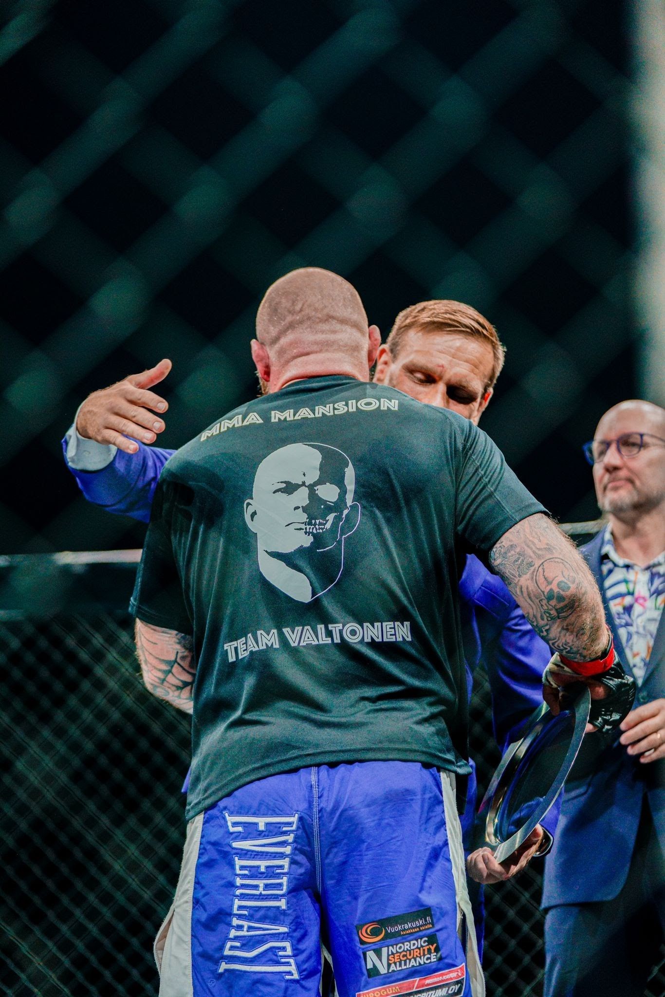 Kaksi miestä ja puvussa oleva toimitsija UFC-häkissä, yksi ottelija takaapäin nähtynä sinisissä shortseissa ja mustassa paidassa