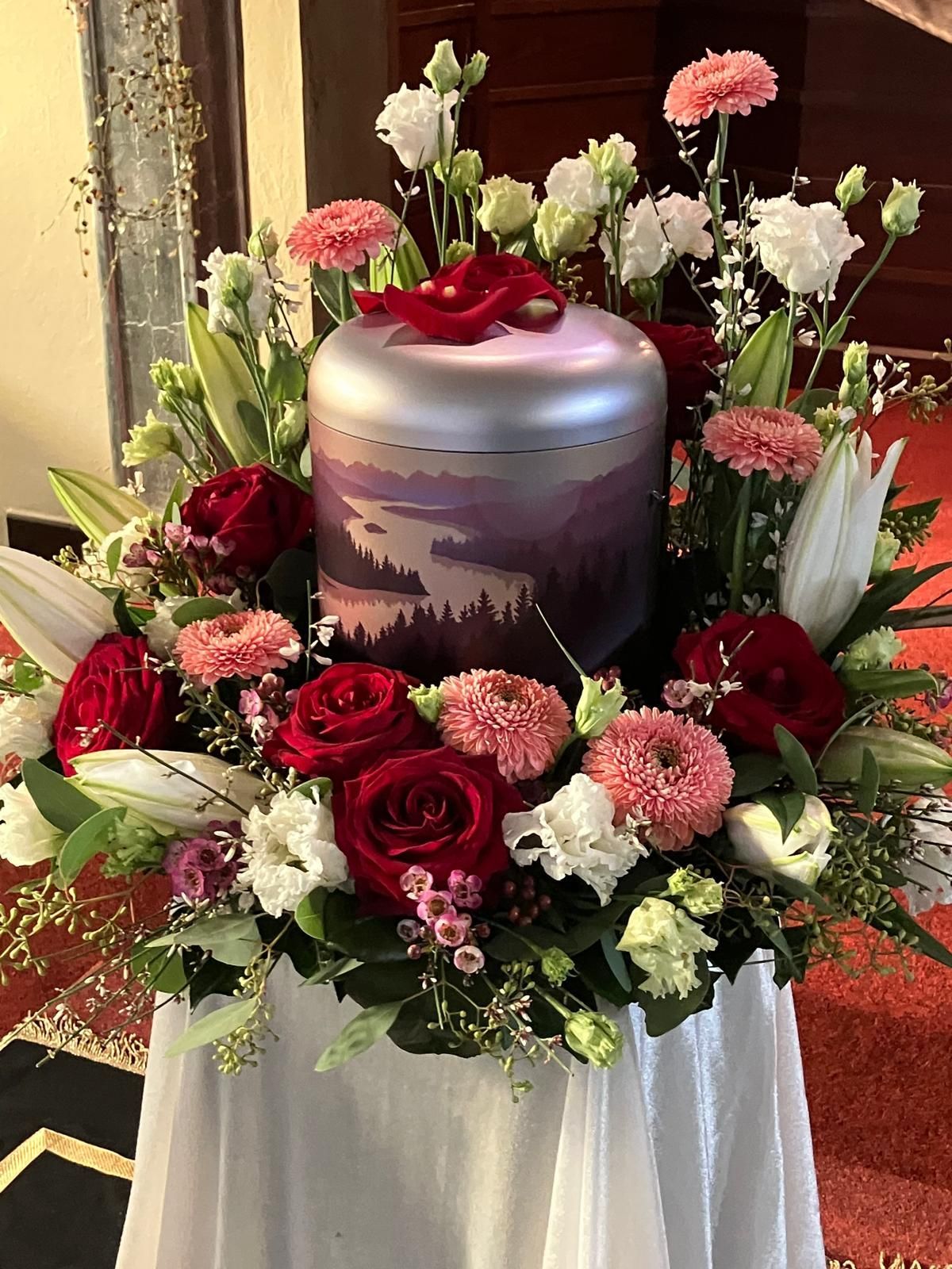 Eine Kremationsurne mit einer Berglandschaftsdarstellung, umgeben von einem Blumenarrangement aus Rosen und Lilien.