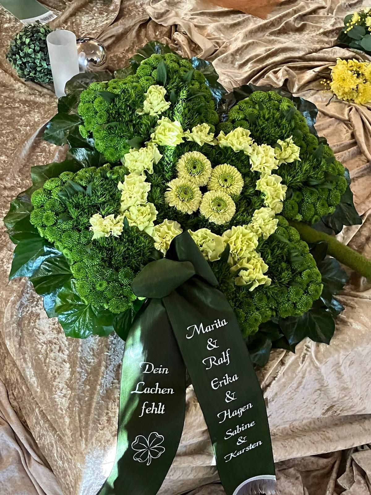 Ein kleeblattförmiges Blumenarrangement auf Stoff mit einem Band, auf dem „Dein Lachen fehlt“ und Namen stehen.