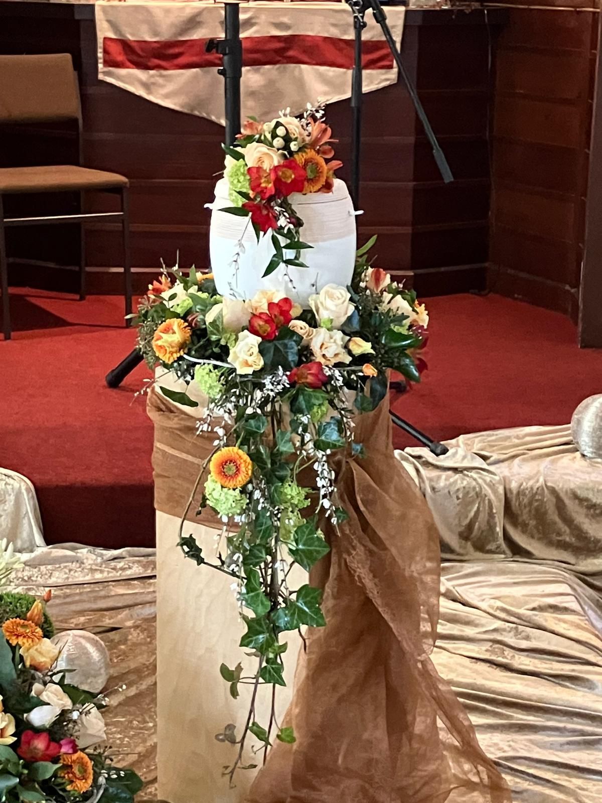 Eine weiße Urnel geschmückt mit einem kaskadenförmigen Blumenarrangement aus orangefarbenen Rosen und grünem Efeu.