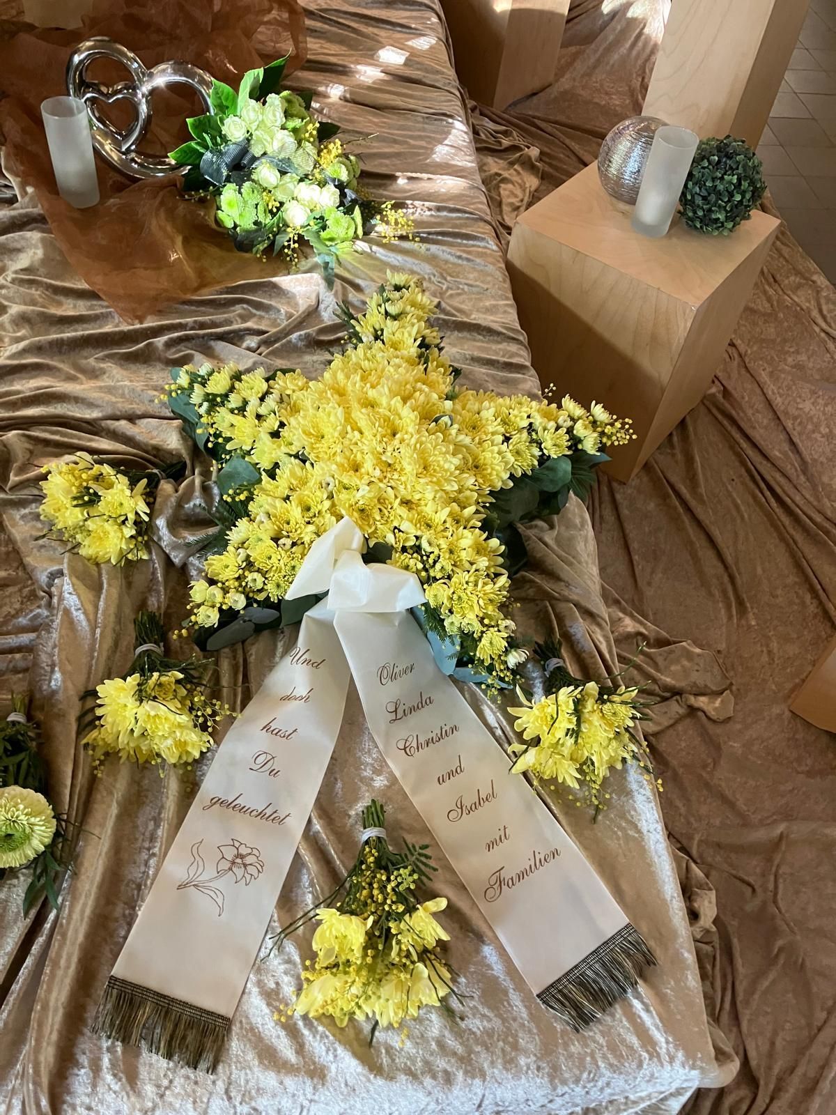 Auf einem Tisch liegt ein gelber Blumenstern als Gedenkgeste, mit zwei weißen Bändern mit handgeschriebenen Inschriften.