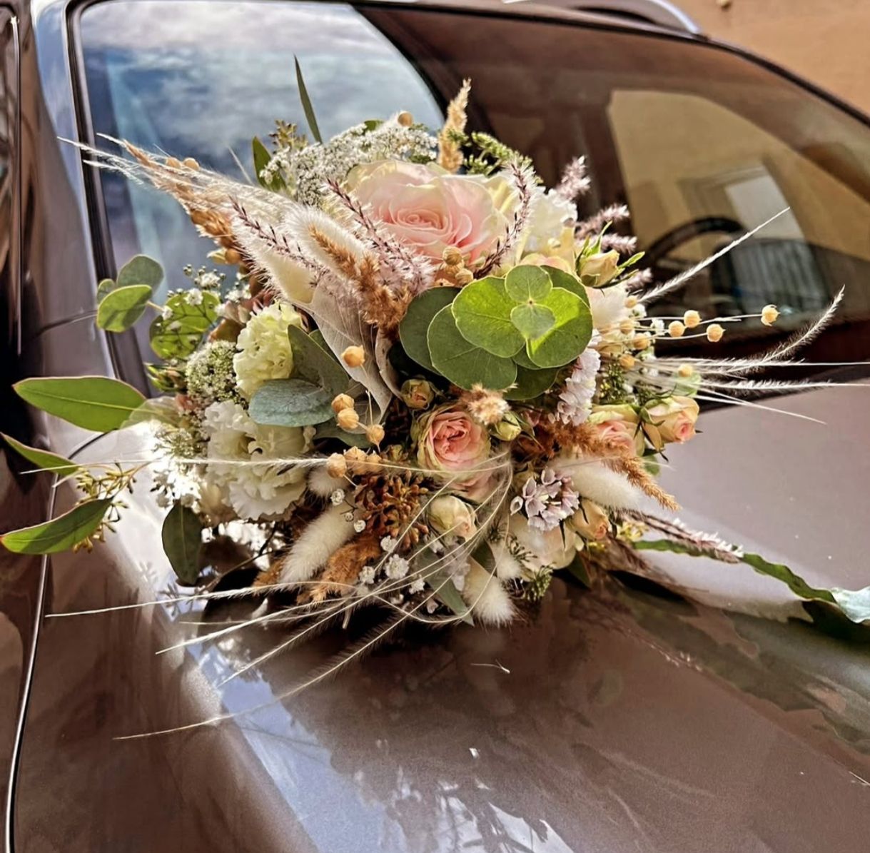 Ein üppiger, rustikaler Strauß aus rosa Rosen, weißen Gänseblümchen, grünem Laub und getrockneten Gräsern.