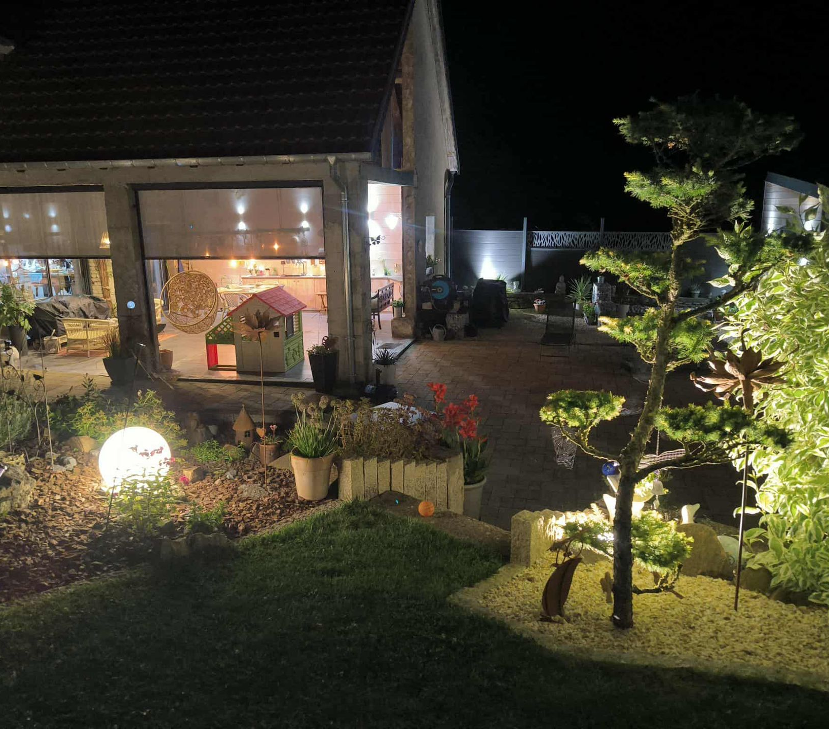 Vue nocturne d'un jardin avec un globe lumineux, un arbre illuminé et un bâtiment aux portes ouvertes.