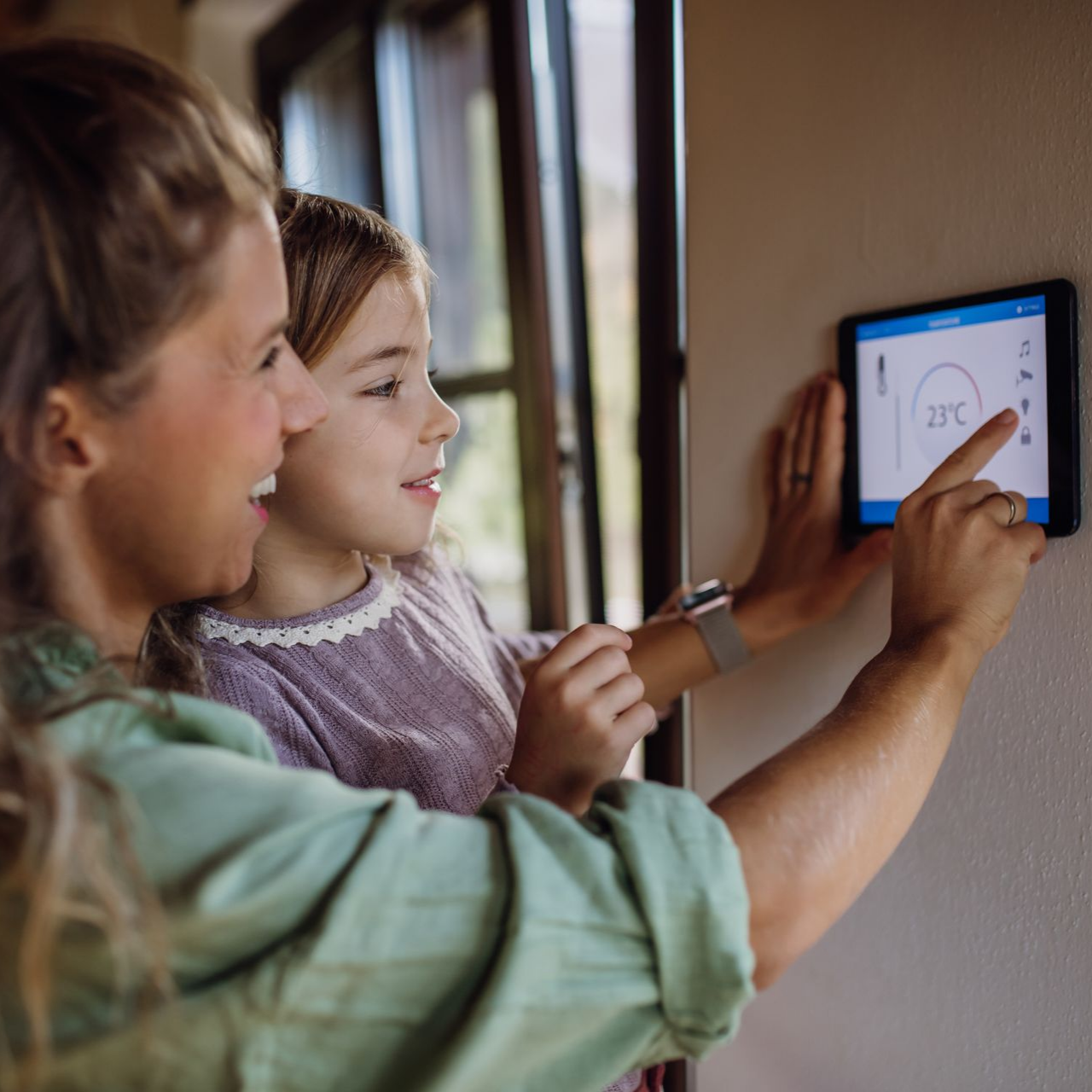 Une femme et un enfant interagissent avec un thermostat intelligent fixé au mur.