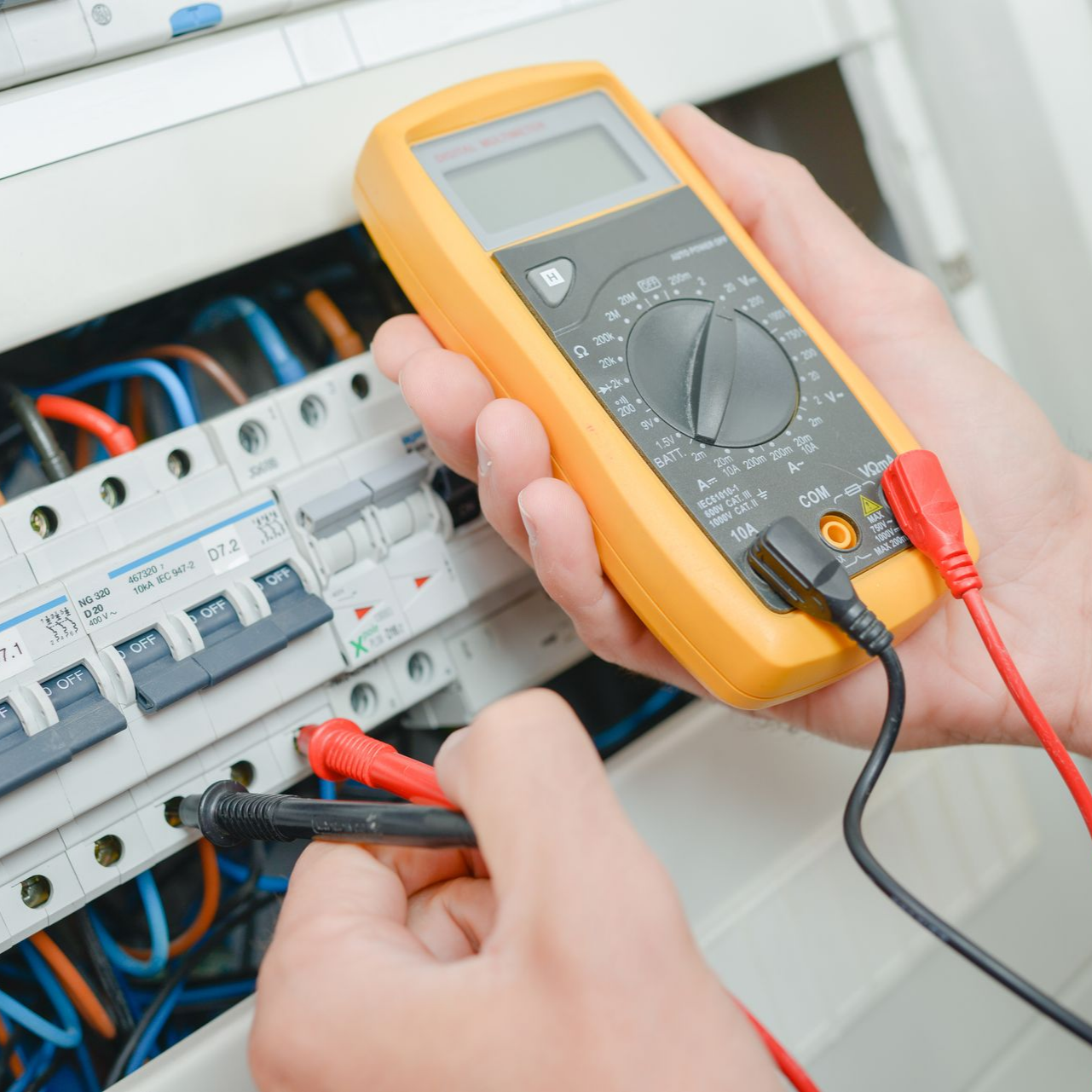 Une personne utilise un multimètre jaune pour tester le câblage électrique d'un tableau électrique.
