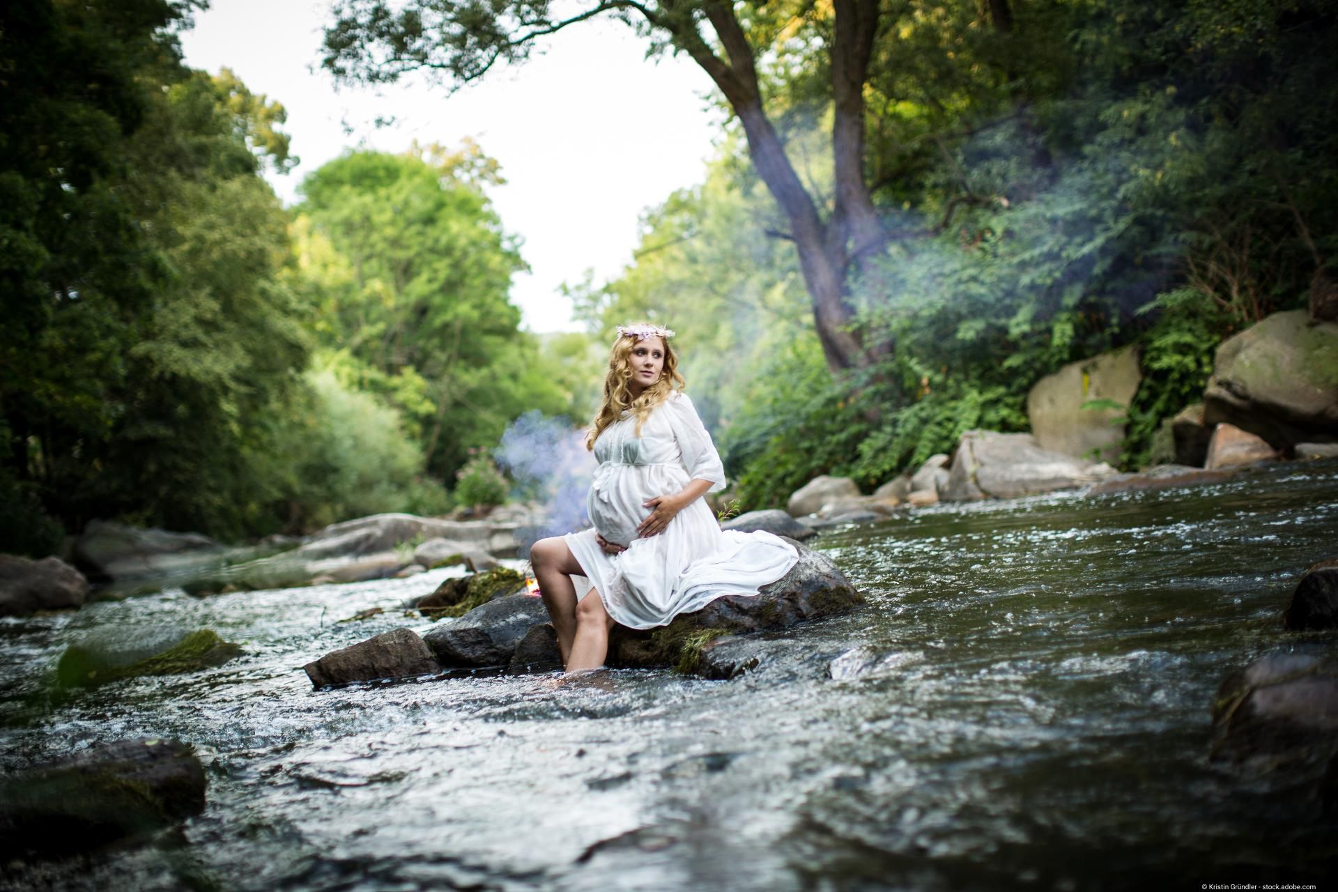 Babybauchfotografie – schwangere Frau sitzt in weißem Kleid am Fluss