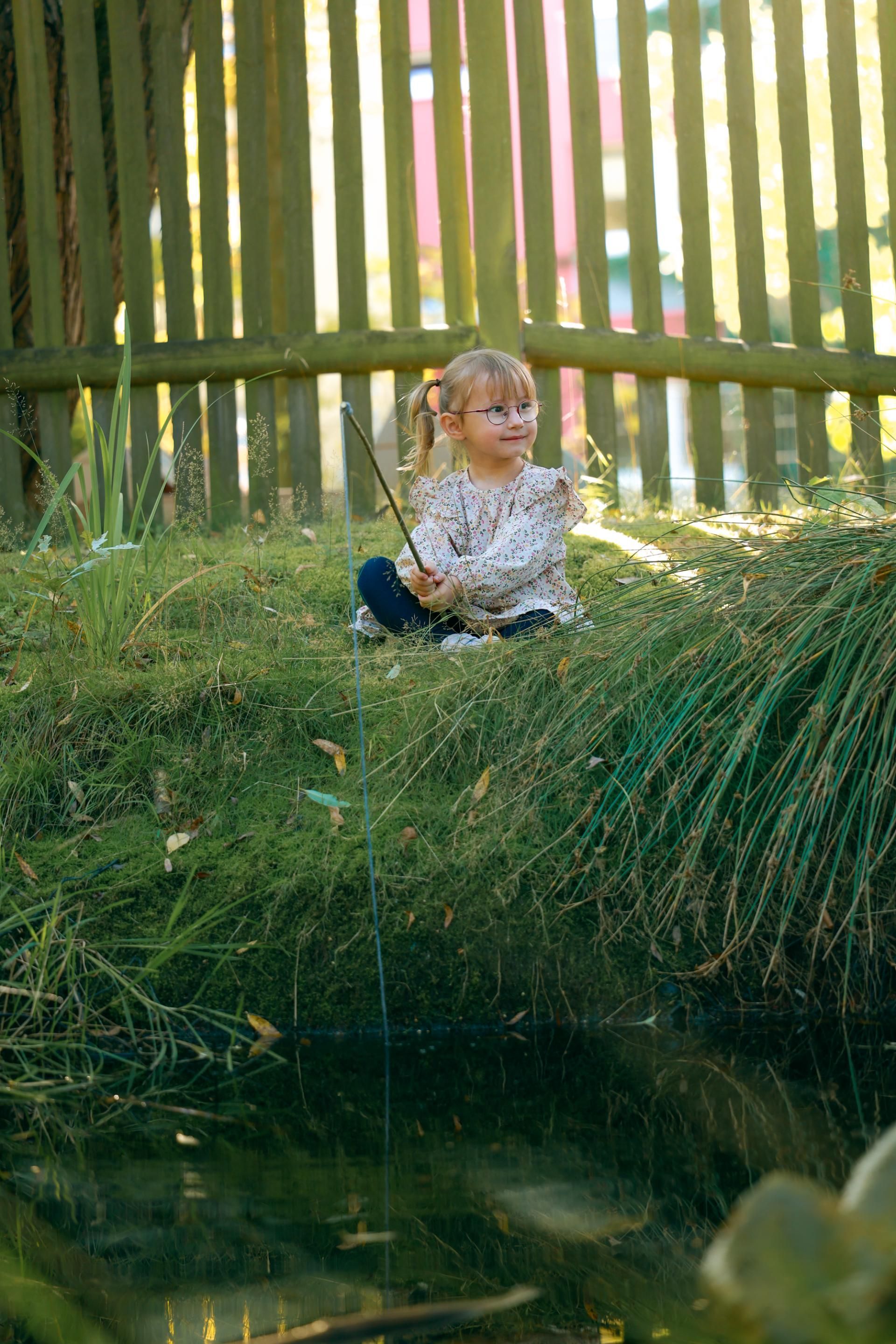 Kinderfotografie – Mädchen sitzt im Gras vor einem Holzzaun